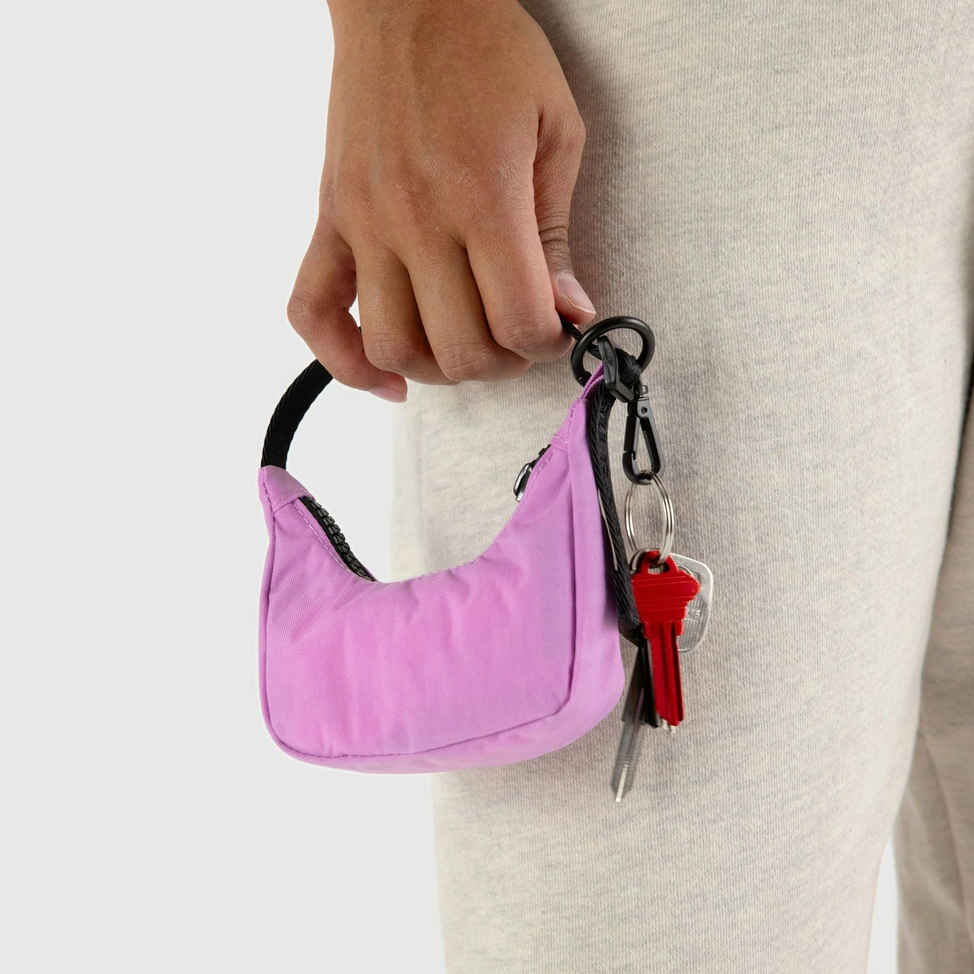 A person holds a small pink handbag, adorned with a Baggu Crescent Bag Charm in Cornflower, and a keyring with keys.