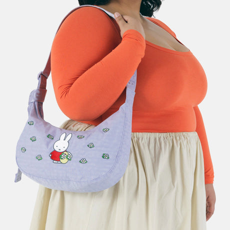A person in an orange top and cream skirt holds the Baggu Miffy x Baggu Medium Nylon Crescent, featuring a cartoon bunny and flowers.