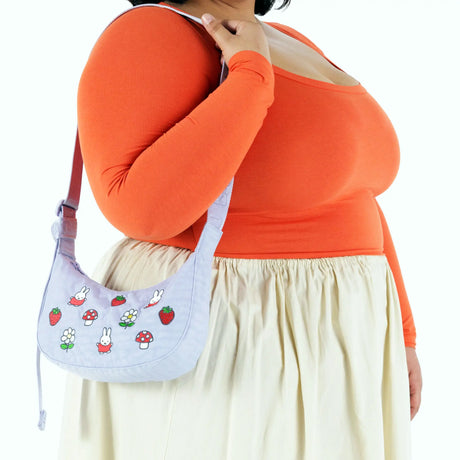 A person in an orange long-sleeve top and cream skirt holds the Baggu Miffy x Baggu Small Nylon Crescent, a lavender bag with cartoon bunnies, strawberries, flowers, and playful Miffy embroidery.