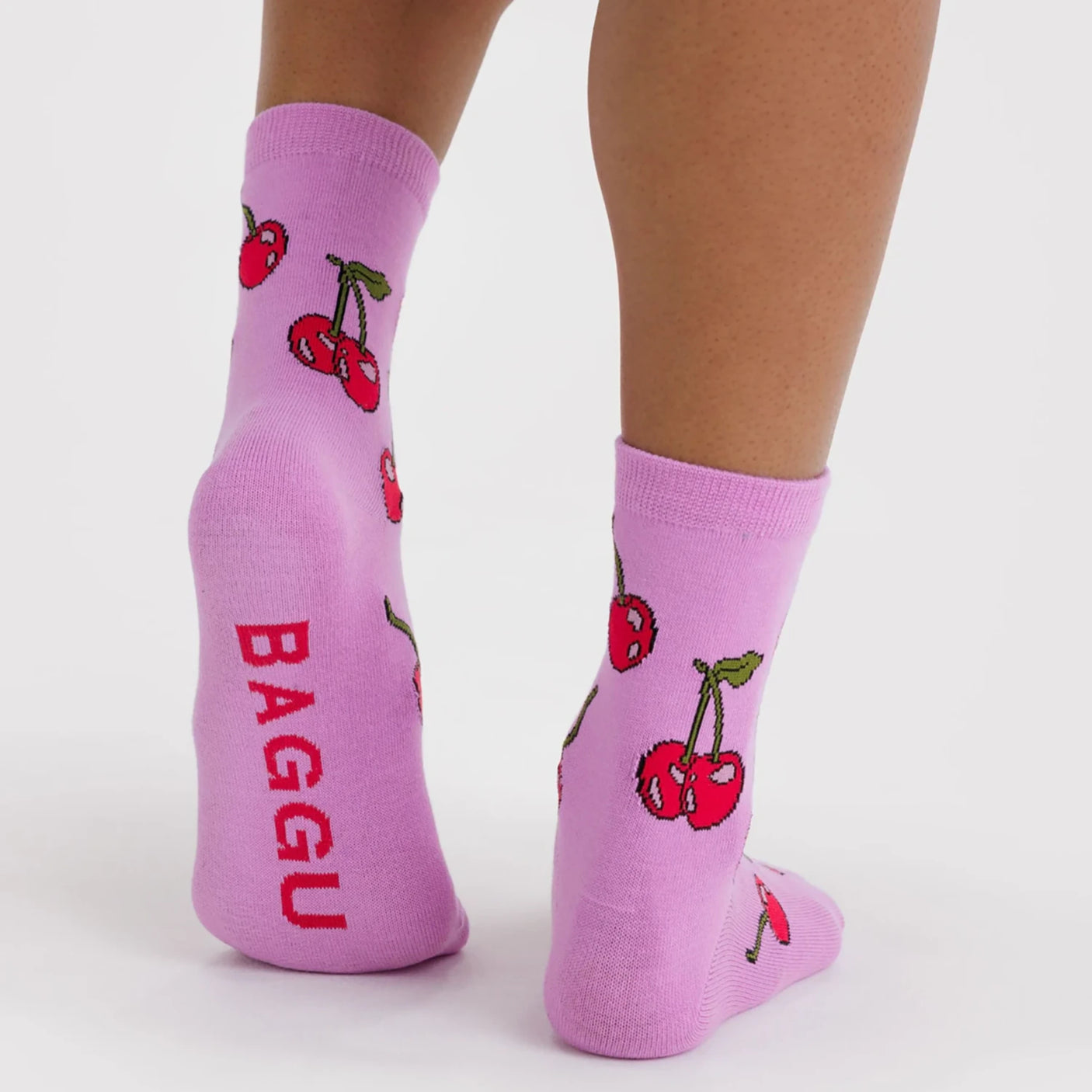 Pair of light purple ankle socks with red cherries and green stems, red text on sole, worn on feet against white background