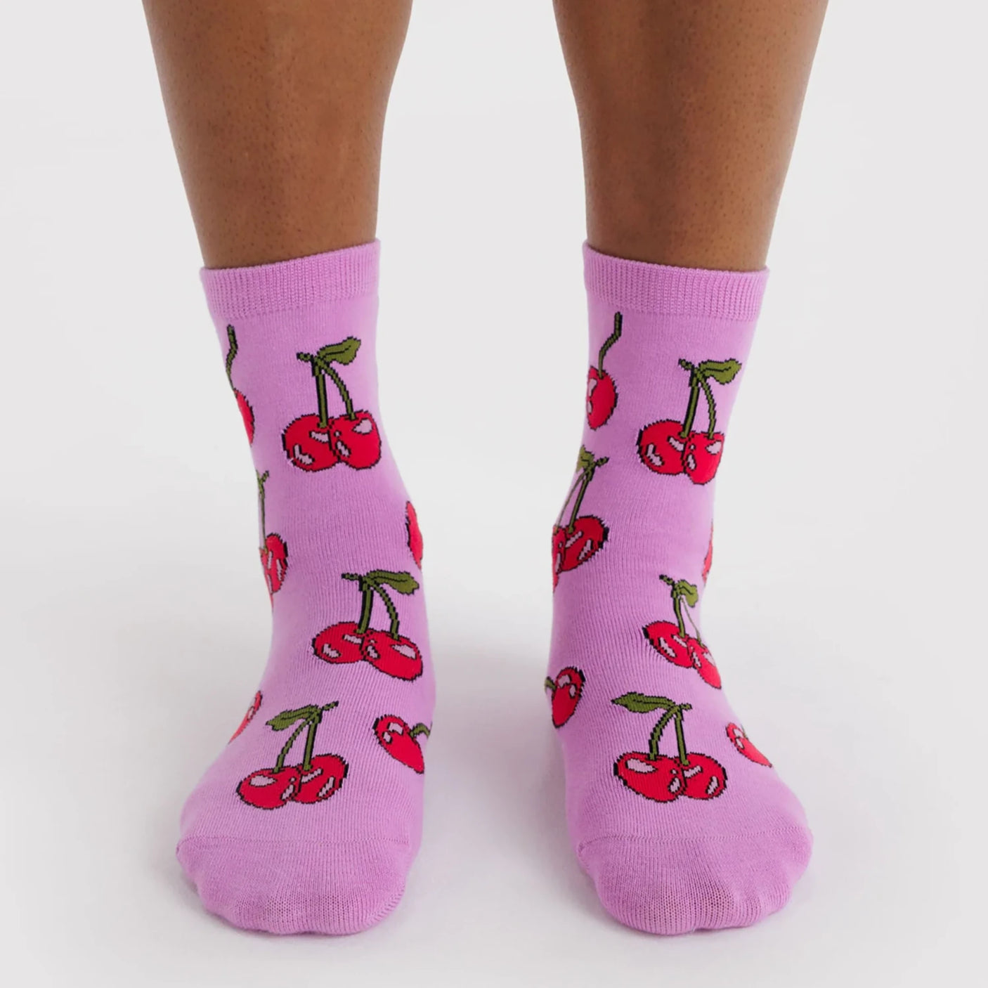 Pair of mid-calf purple socks featuring red cherries with green stems worn on feet against a white background