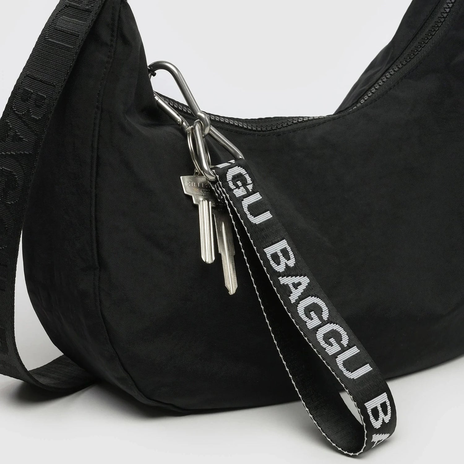 A black shoulder bag features a BAGGU-branded strap of recycled nylon webbing with a pair of keys attached by a sturdy metal clip to the Baggu Logo Keychain - Peony.