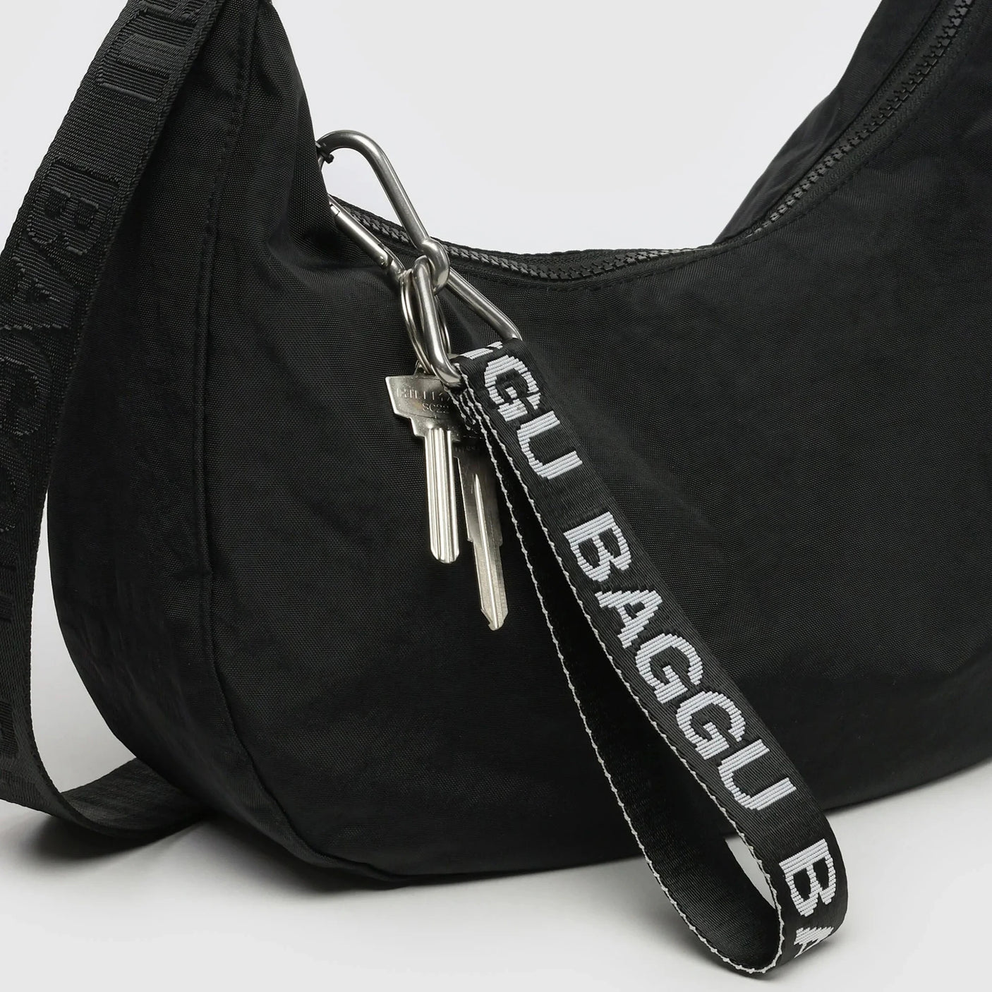 A black shoulder bag features a BAGGU-branded strap of recycled nylon webbing with a pair of keys attached by a sturdy metal clip to the Baggu Logo Keychain - Peony.