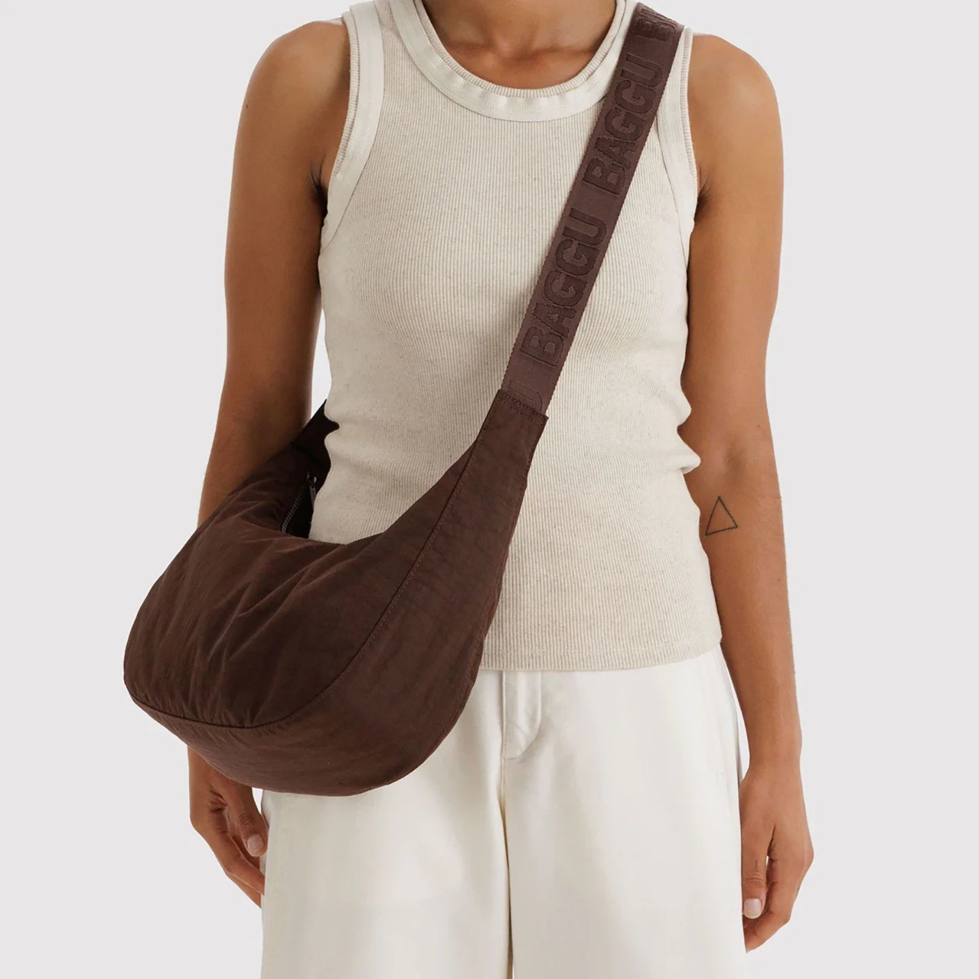 Chocolate brown crescent-shaped fabric crossbody bag with wide branded strap worn by person in beige tank top and white pants