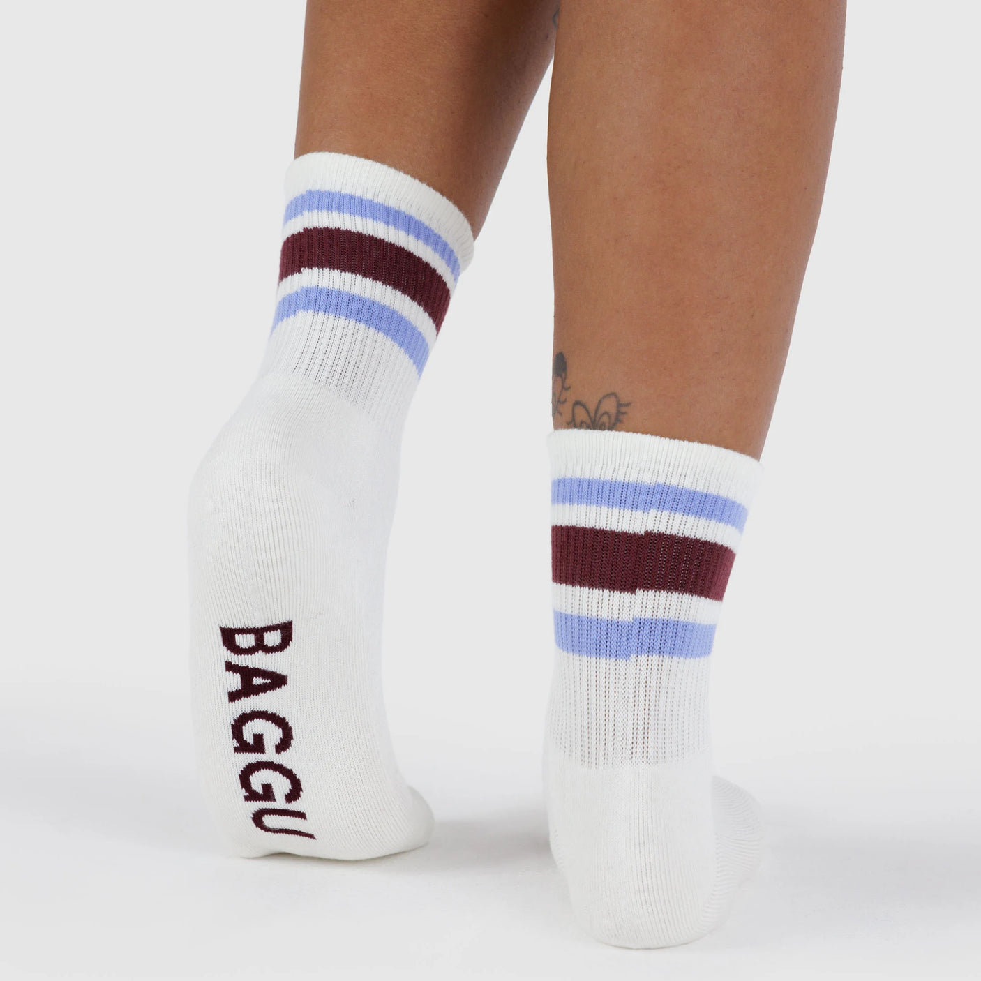 A person stands on a white background wearing Baggu Ribbed Sock - Ivory Stripe with blue and maroon stripes and “BAGGU” on the sole. A partial tattoo is visible, drawing attention to these classic striped socks by Baggu.