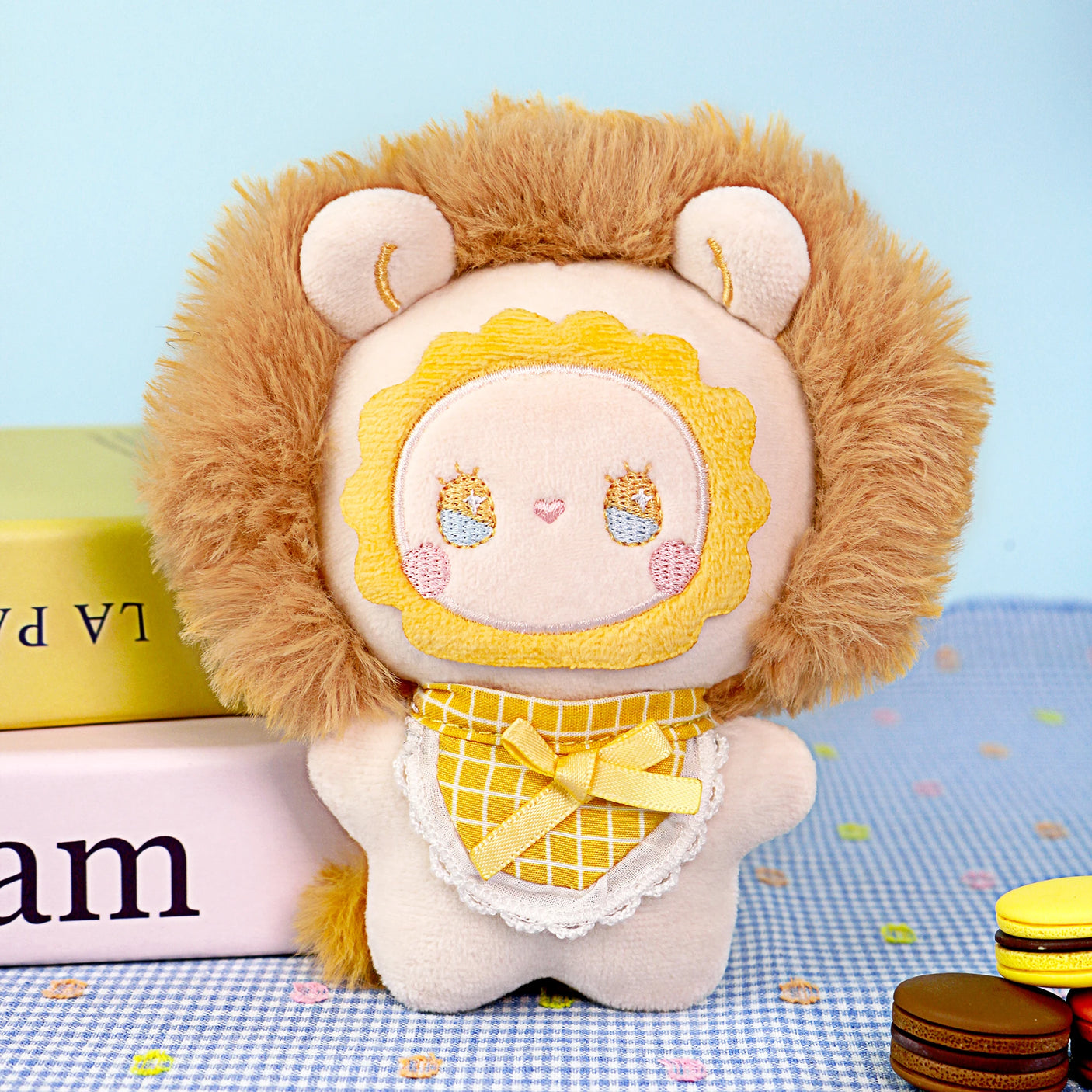 Discover the Lucky Emma Fuzzy Family Keychain Blind Box: a plush lion toy with a fluffy mane and yellow checkered bib, presented on a blue dotted surface beside stacked books and toy macarons—an adorable bag accessory.