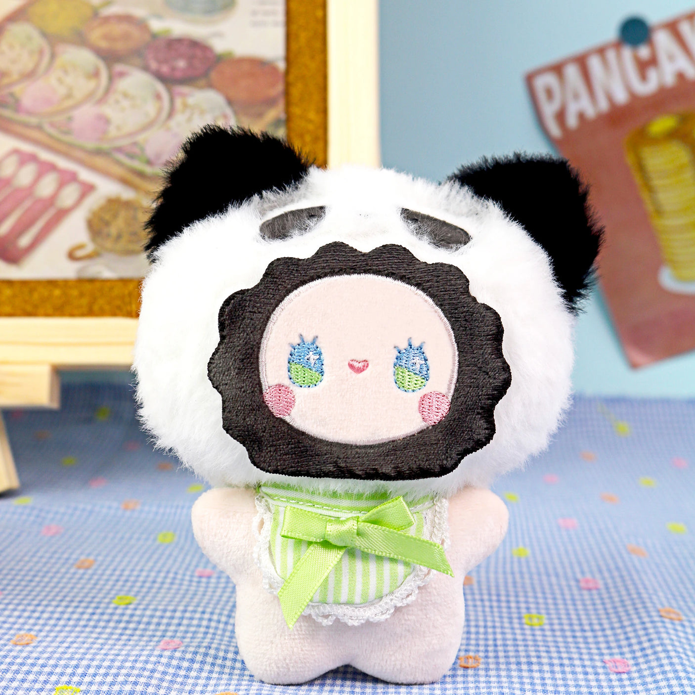 The Lucky Emma Emma Fuzzy Family Keychain Blind Box features a plush doll with a panda hood, green ribbon, and embroidered face on a blue polka-dotted surface—ideal as a cute keychain or bag charm. Blurred background decor adds charm.