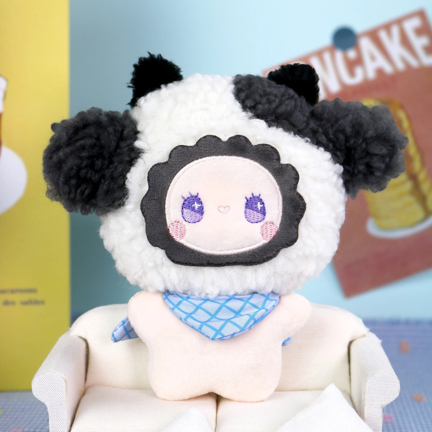 The Emma Fuzzy Family Keychain Blind Box by Lucky Emma features a plush doll in a sheep costume with black ears and a blue scarf, seated on a white sofaβideal as a keychain or bag charm. Bright wall dΓ©cor enhances the sceneβs cute appeal.