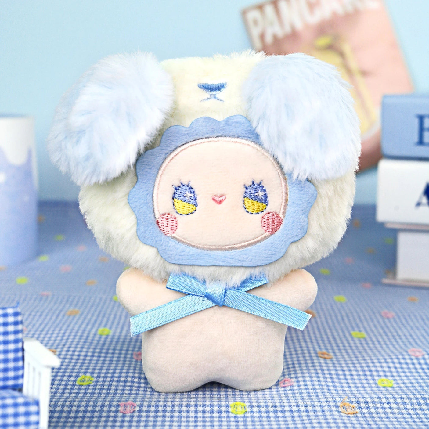 The Lucky Emma "Emma Fuzzy Family Keychain Blind Box" features a plush toy with a round embroidered face, blue ears, and a blue ribbonβperfect as a cute keychain or bag charm. Books and soft props set the scene in the background.