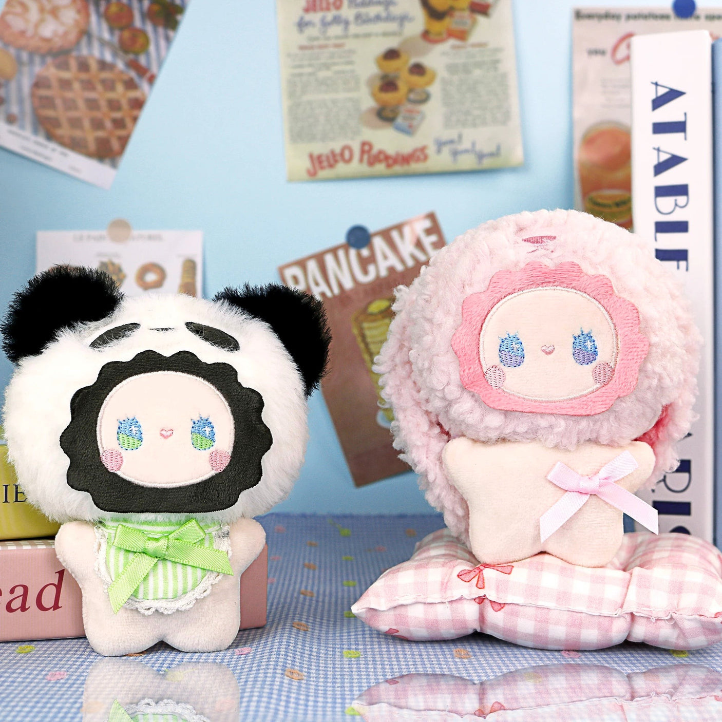 Two pastel plush dolls with round faces are shown on a dotted surface; one wears a panda hat like the Lucky Emma Fuzzy Family Bag Charm - Blind Box, while the other has a fluffy pink hood and sits on a checkered pillowβgreat as animal bag charms.