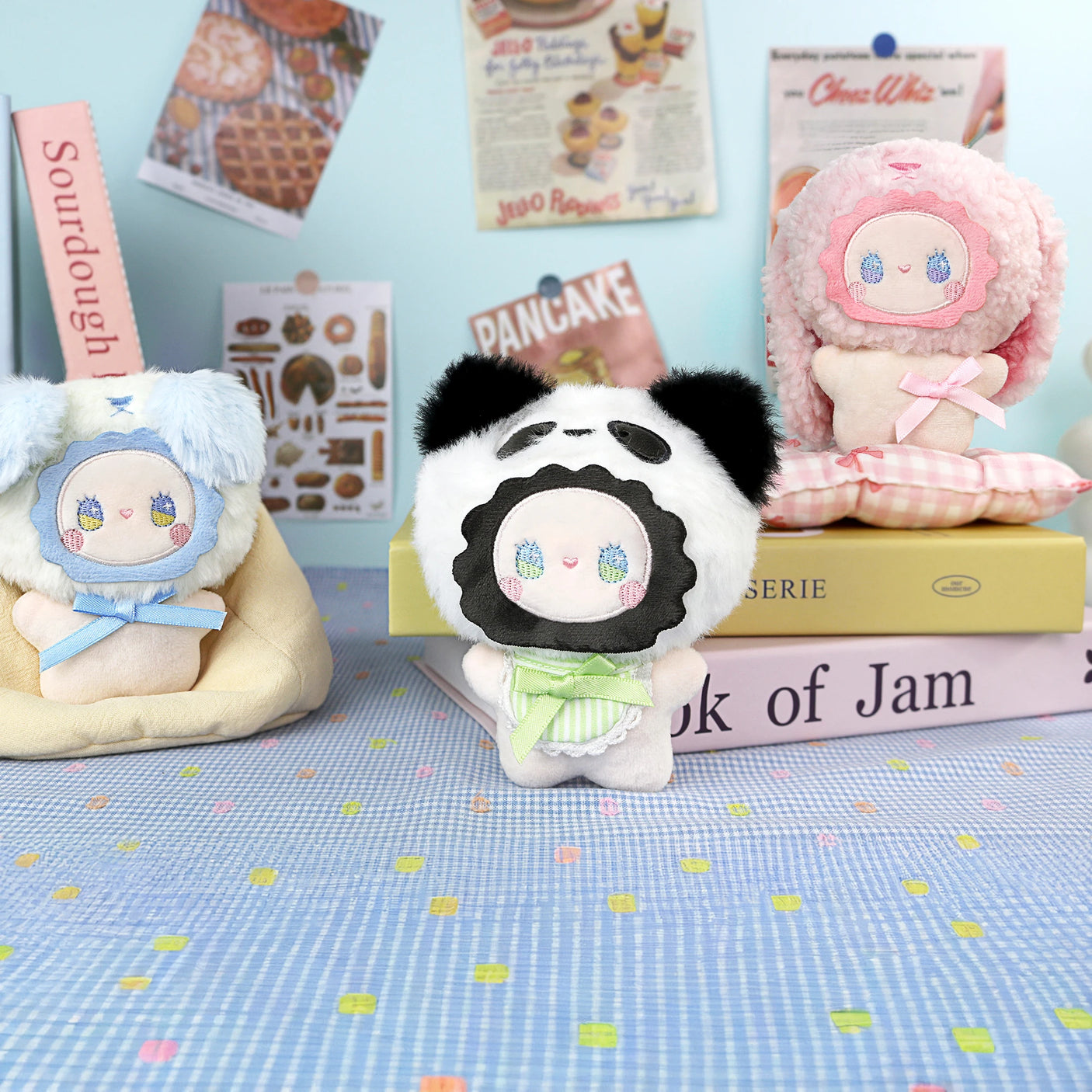 Three Emma Fuzzy Family plush dolls by Lucky Emma, each with animal-themed hoods and pastel colors, are displayed on a table with books as food-themed posters decorate the backdrop; each doubles as a plush keychain for your bag.
