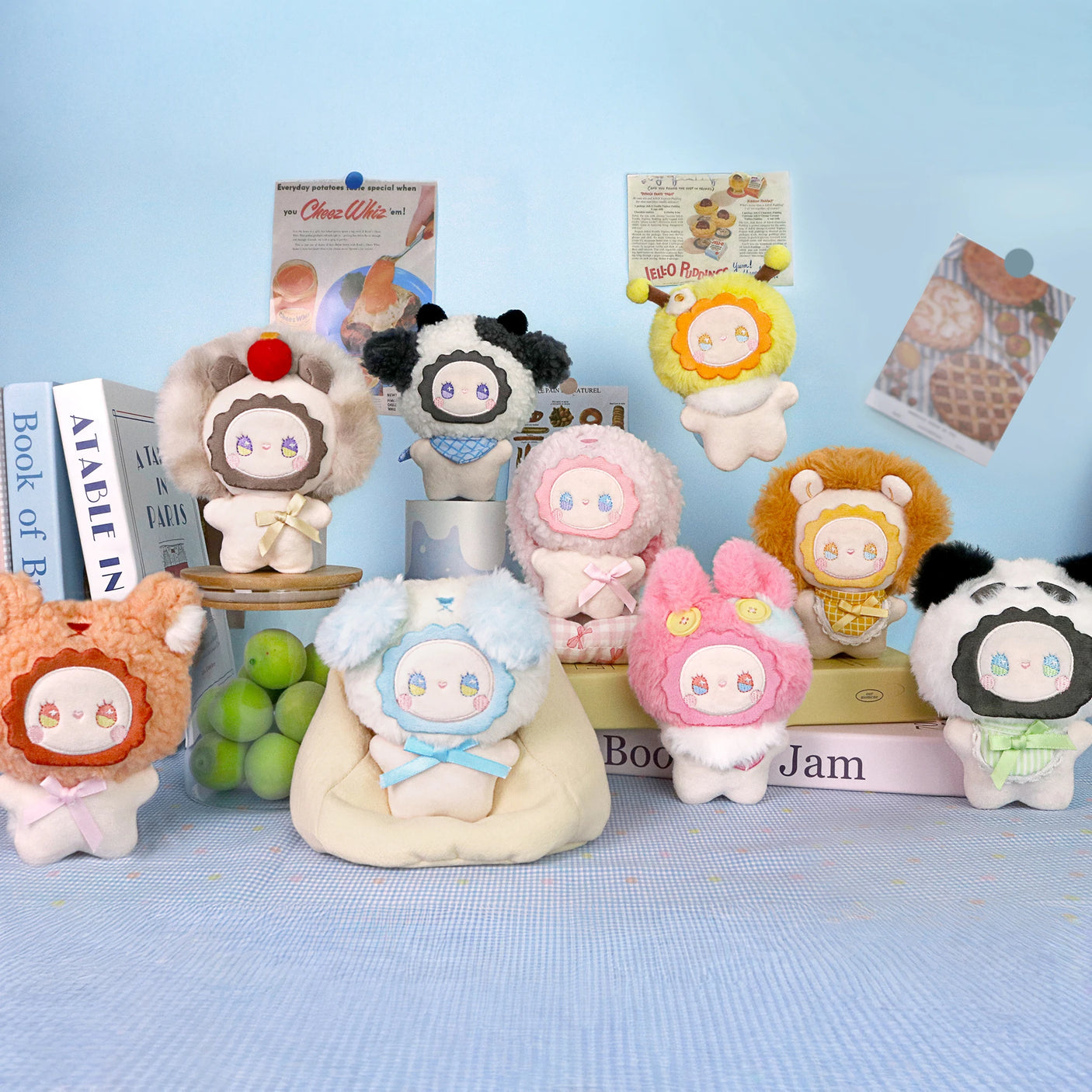 Ten pastel animal-themed plush toys from Lucky Emma are displayed on books, showcasing the Emma Fuzzy Family Bag Charm - Blind Box, with grape and food props in the background.