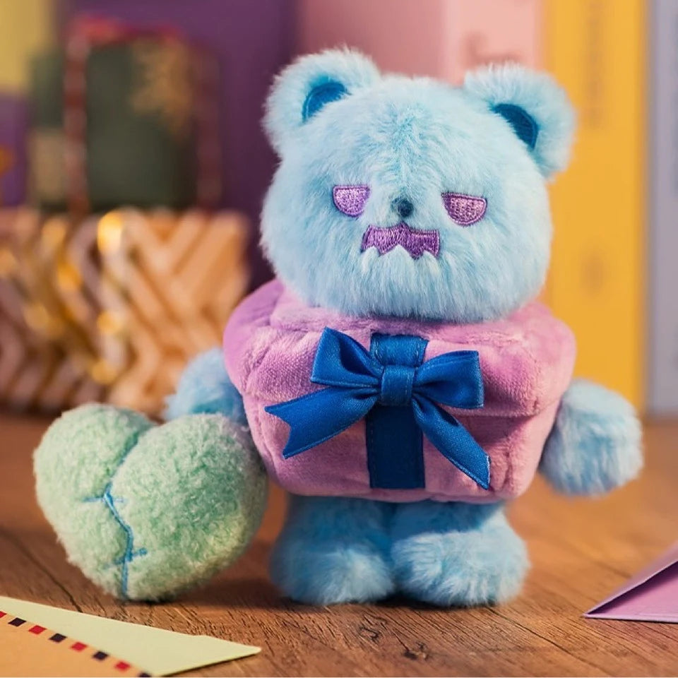 Blue plush bear with purple sleepy eyes wearing a pink gift box outfit and holding a green heart plush on wooden surface