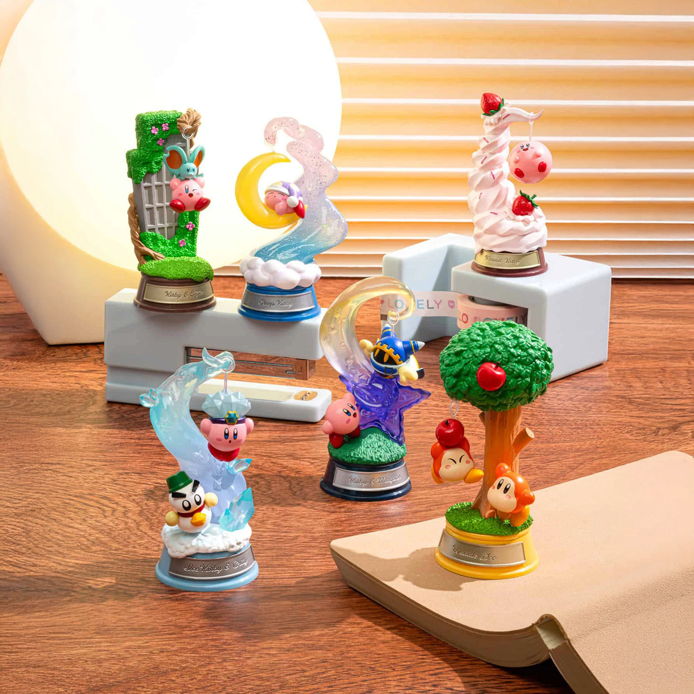 Six vibrant Kirby figures from the Kirby Dream Land Blind Box by Kirby are displayed on a wooden surface near a lamp and a closed book, each showcasing different characters and scenes from the series.