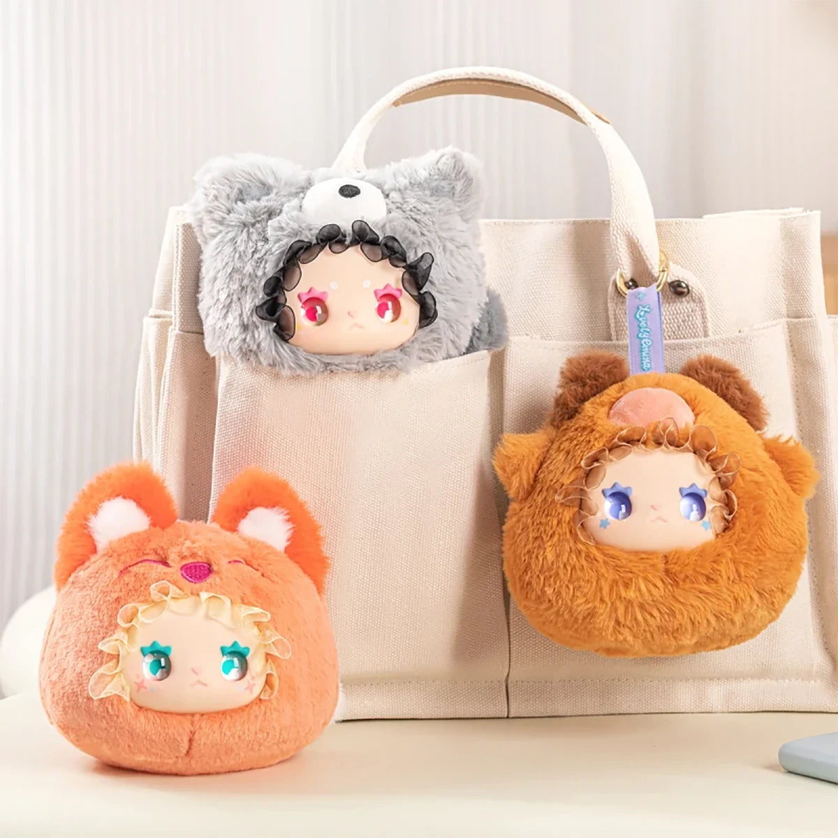 Three Lovely Emma Rua Rua Zoo plush bag charms in fuzzy animal costumes displayed on and near beige tote bag