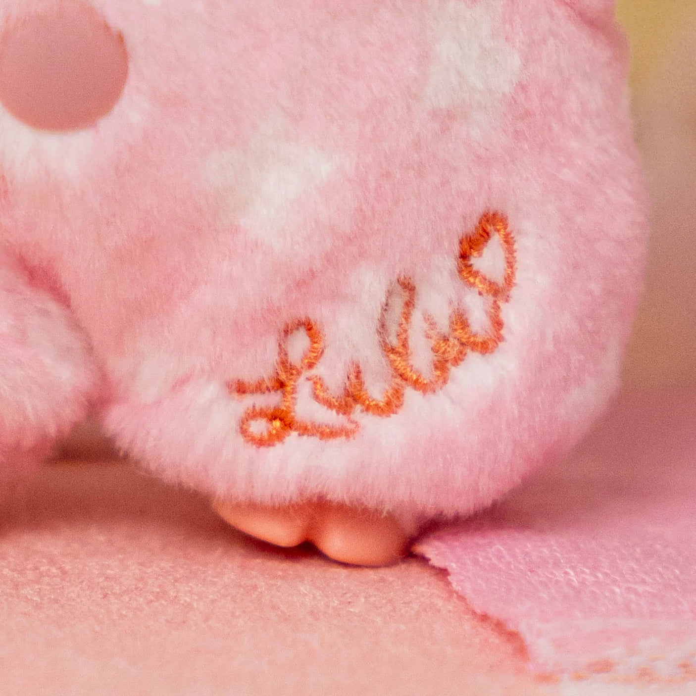 Close-up of pink plush fabric with embroidered "Lulu" signature and tiny plush feet on soft textured surface