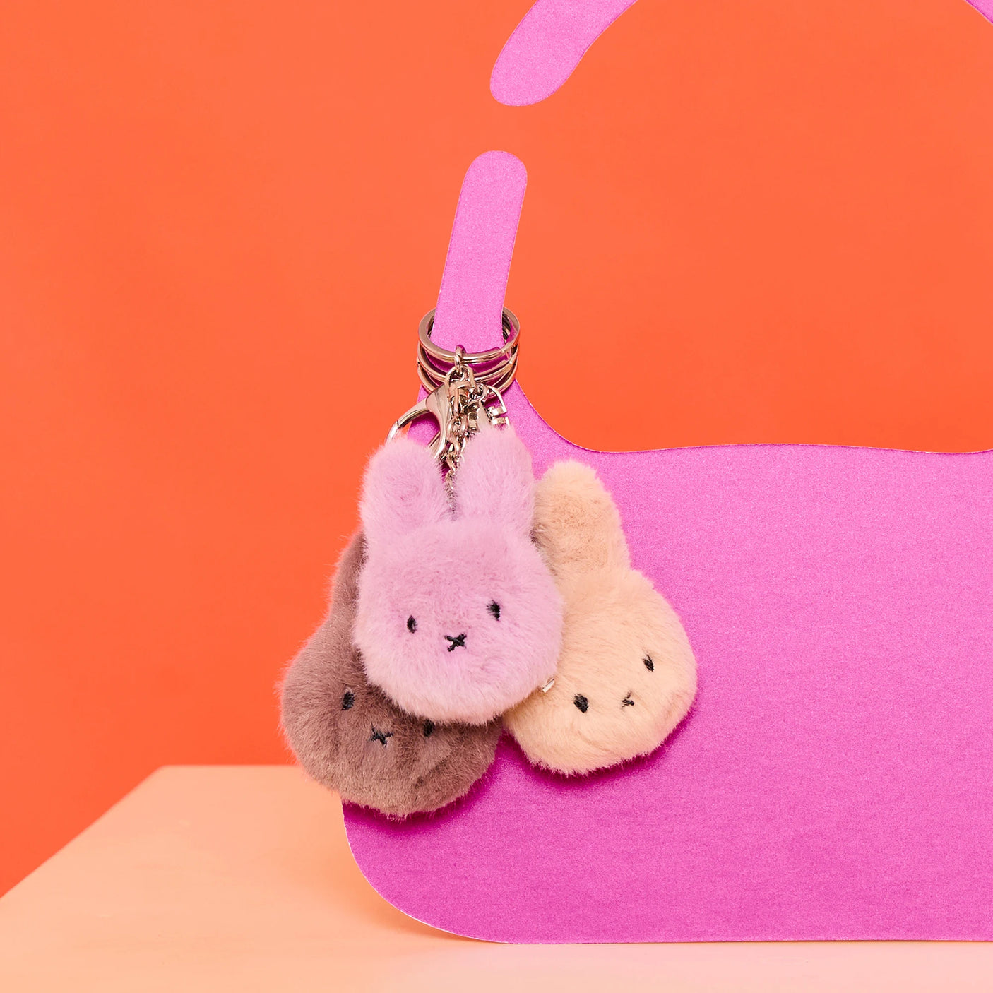 The Miffy Fluffy Keychain in Beige by Miffy hangs on a pink handbag against an orange background, adding a cute, eco-friendly touch with its plush recycled polyester design.