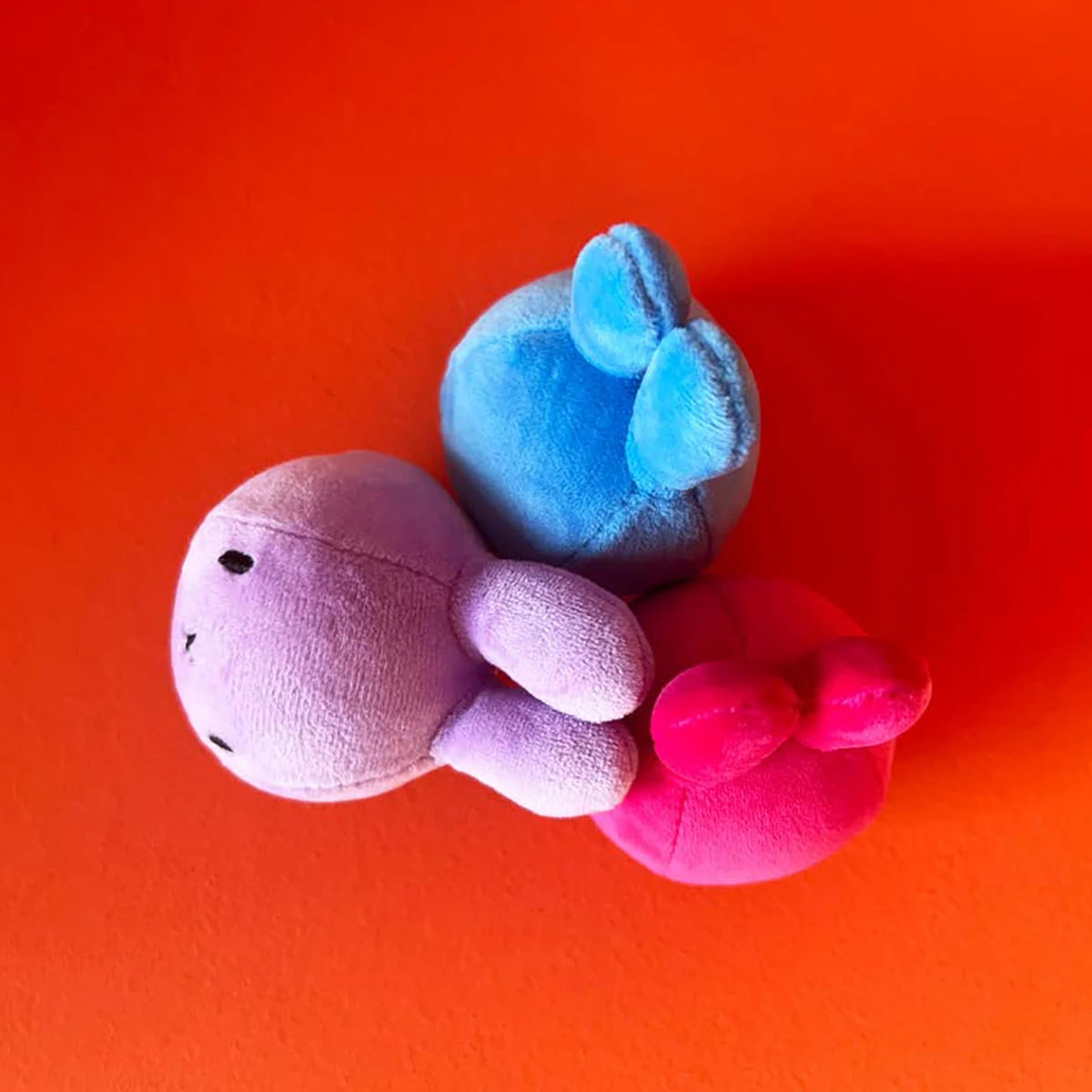 Three Miffy Squish Relief Balls in lilac from Miffy are arranged on an orange surface, their heads facing outward. Each features eco-friendly filling for a soft, stress-busting cuddle experience.