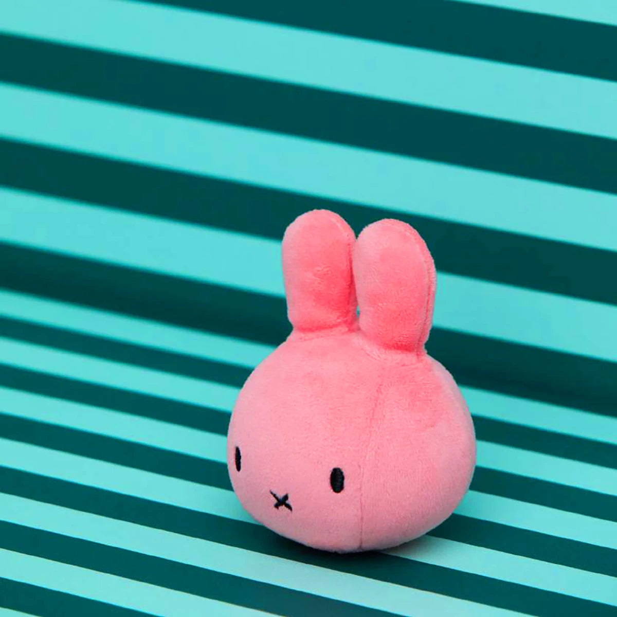 The Miffy Squish Relief Ball - Pink by Miffy is a small, round sensory toy with bunny ears and a simple face, ideal for stress relief. It rests on a teal and dark green striped surface—a cute choice for soothing fun.