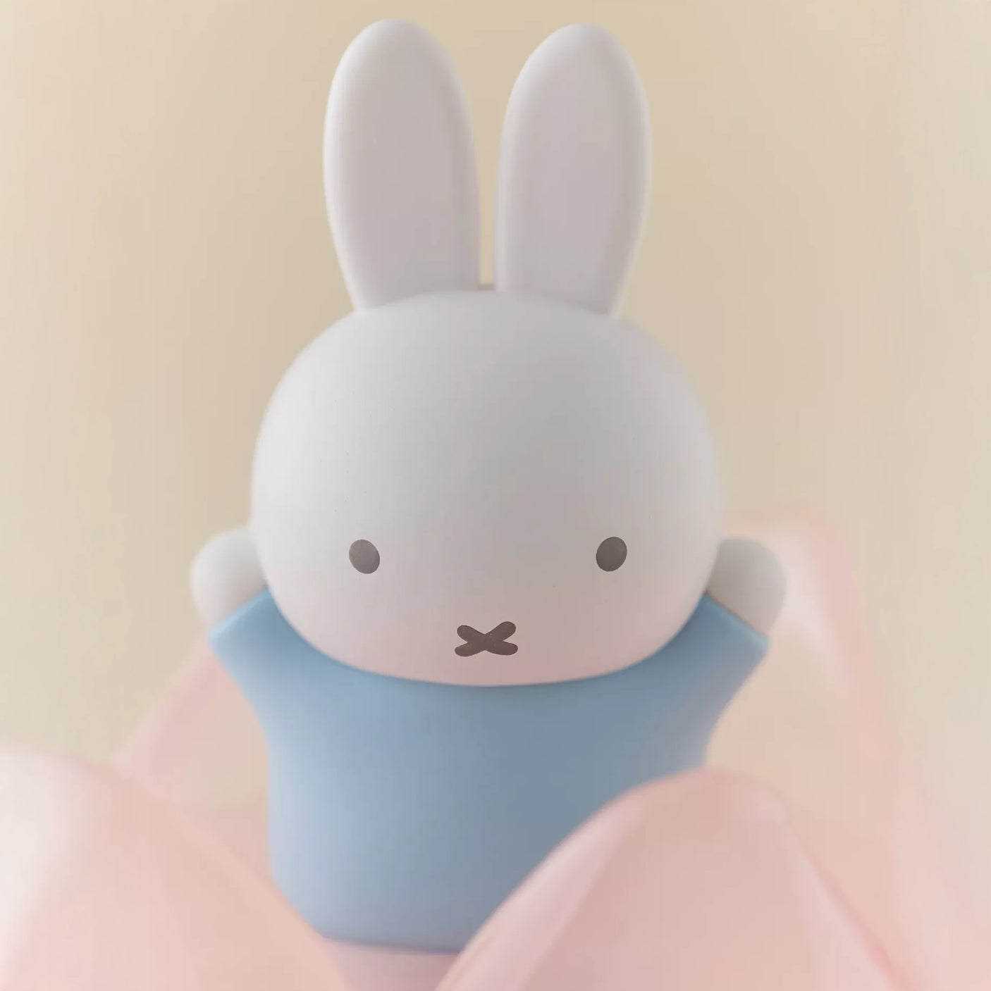 Miffy figure in light blue outfit with white ears surrounded by soft pink fabric on beige background