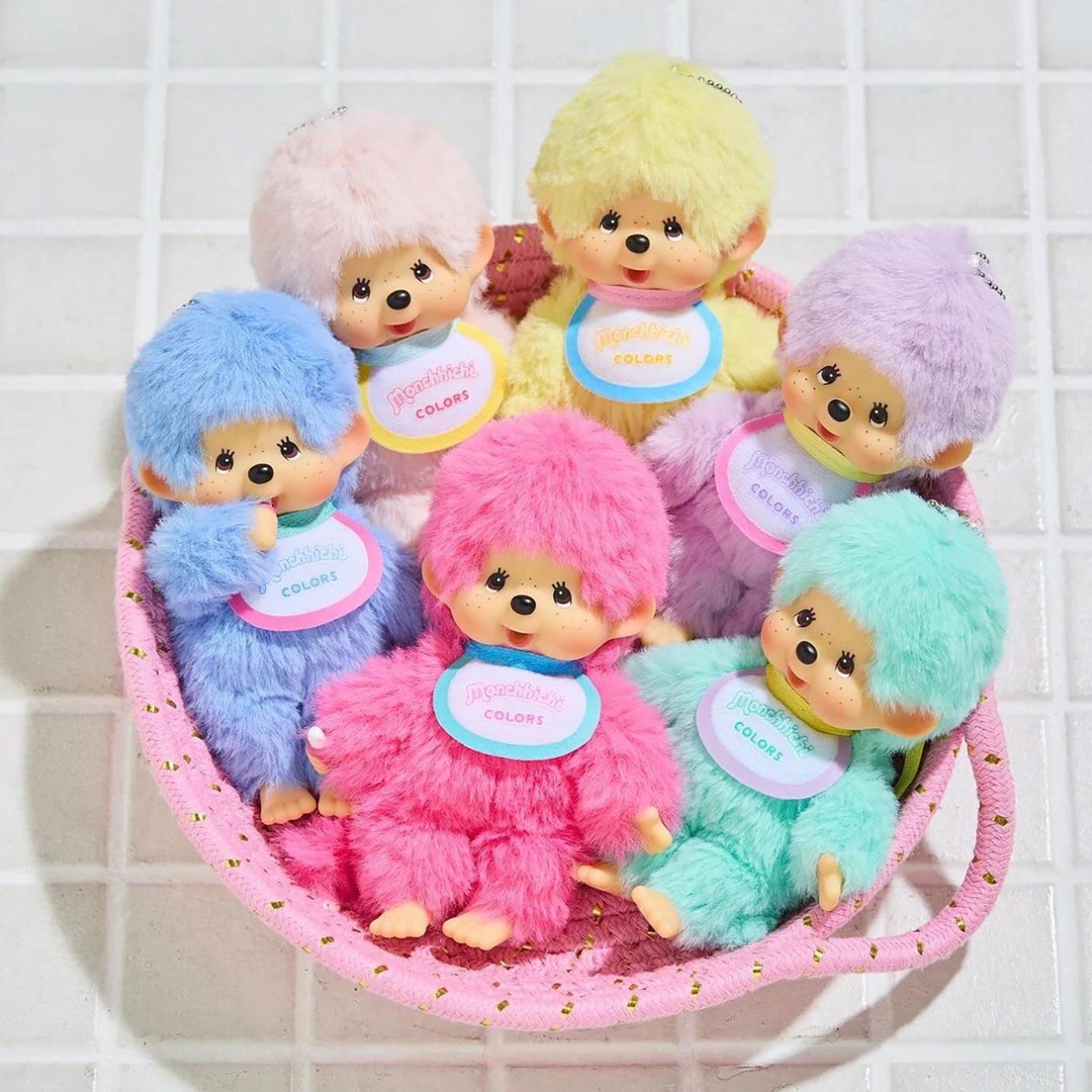 The Monchhichi Colours Bag Charm - Blue by Monchhichi features a plush doll with a round face, fluffy fur, and bib, perfect as a keychain or bag charm gift. Shown in a pink basket with pastel variants on a tiled surface.