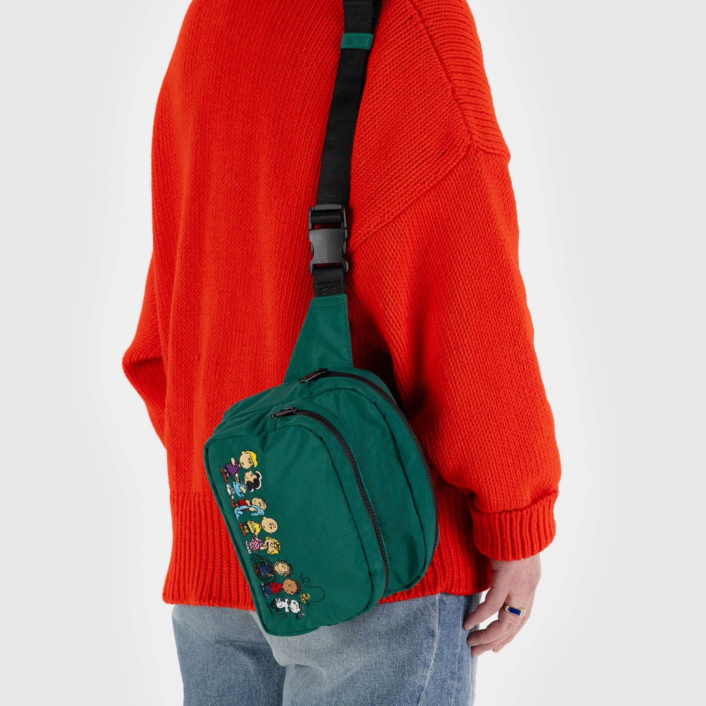 A person wearing a bright orange sweater and blue jeans carries the limited edition Peanuts x Baggu Fanny Pack crafted from recycled nylon by Baggu.