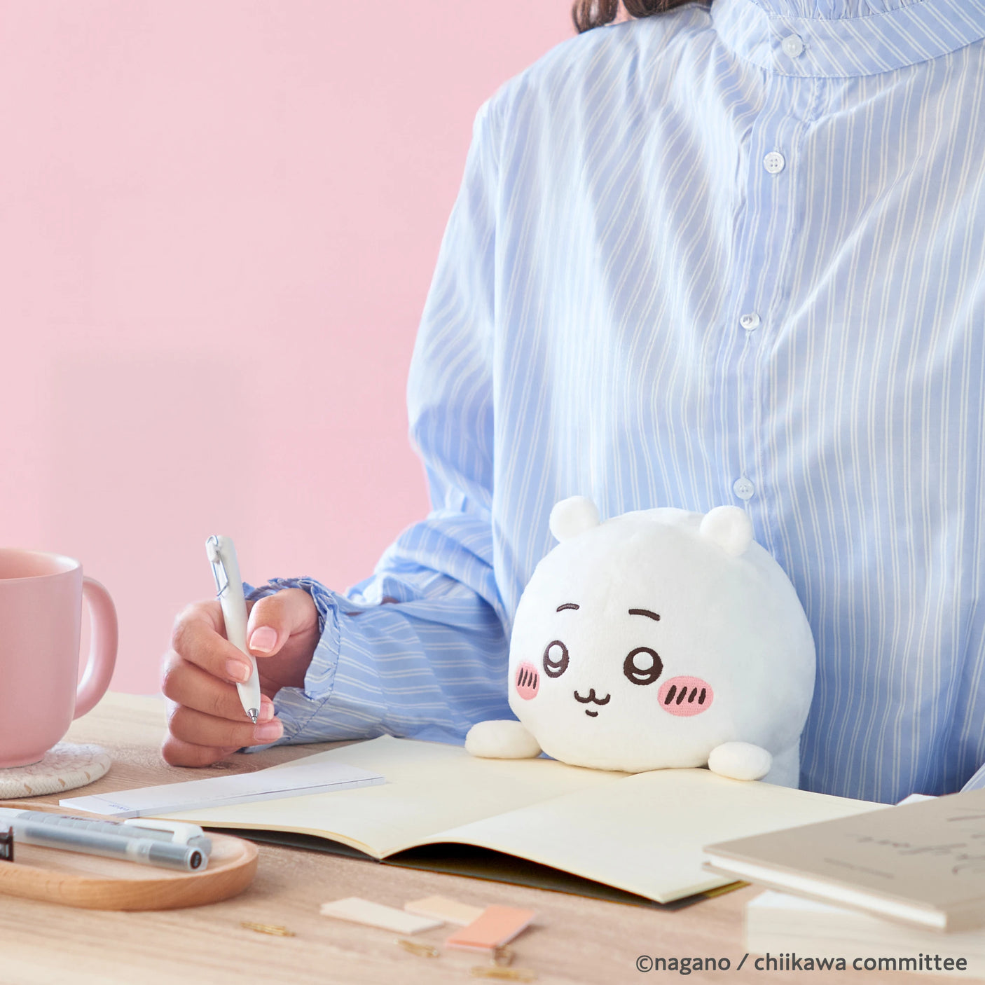 A person in a blue striped shirt writes at their desk with the Chiikawa Posture Plush Buddy by Chiikawa, a pink mug, and stationery—the ideal companion for cozy study sessions.