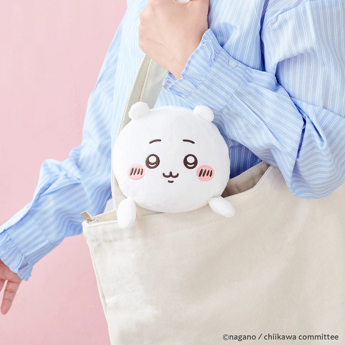 A person in a blue striped shirt carries a beige tote bag with the Chiikawa Posture Plush Buddy by Chiikawa—a round, white plush toy with a cute face and blushing cheeks—peeking out. An adorable companion for your desk!.