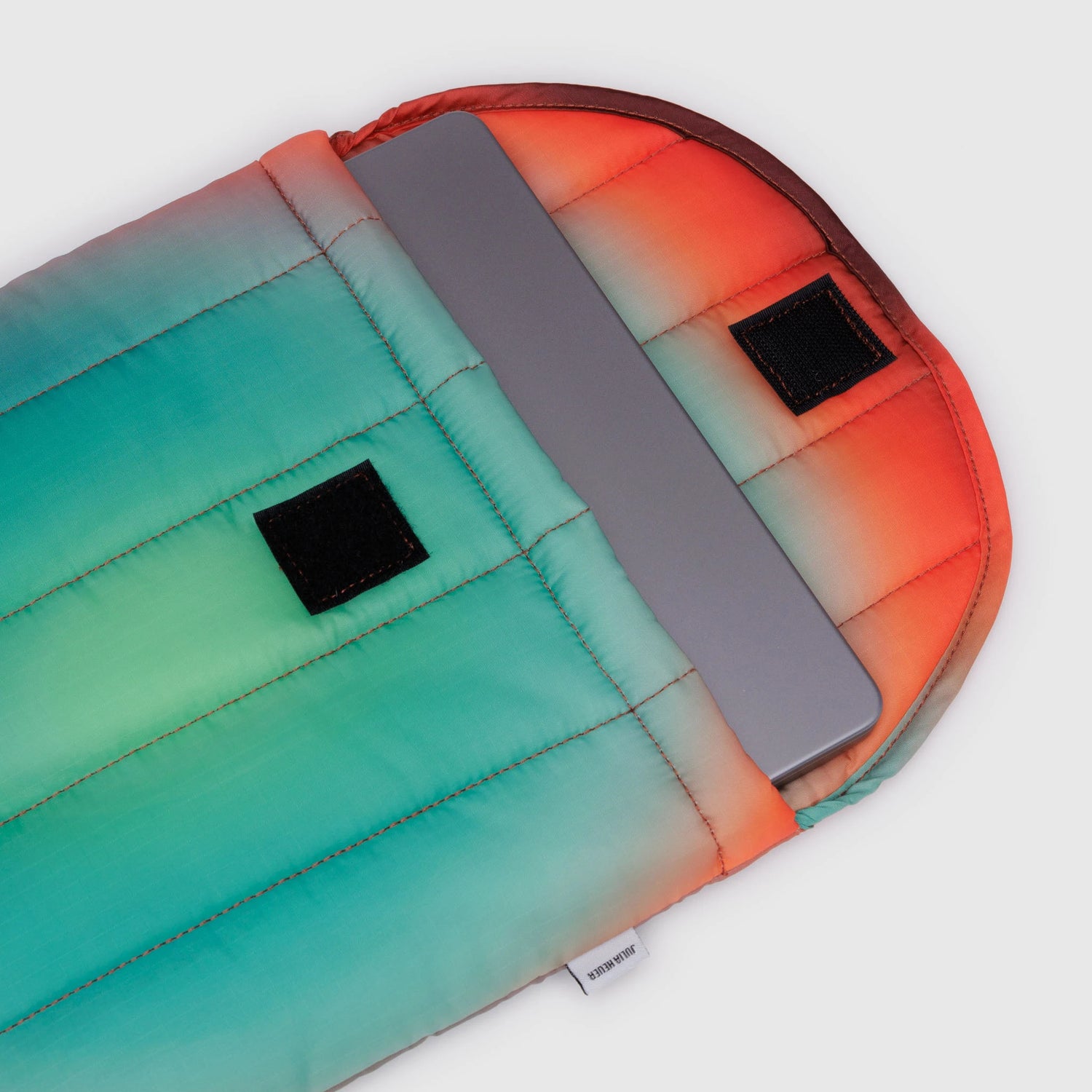 A laptop partially nestled in a colorful, padded sleeve featuring gradient shades of orange, red, teal, and green. This Baggu Puffy Laptop Sleeve 13"/14" - Rain boasts two sleek black Velcro strips on the flap—a true Gradient Masterpiece.