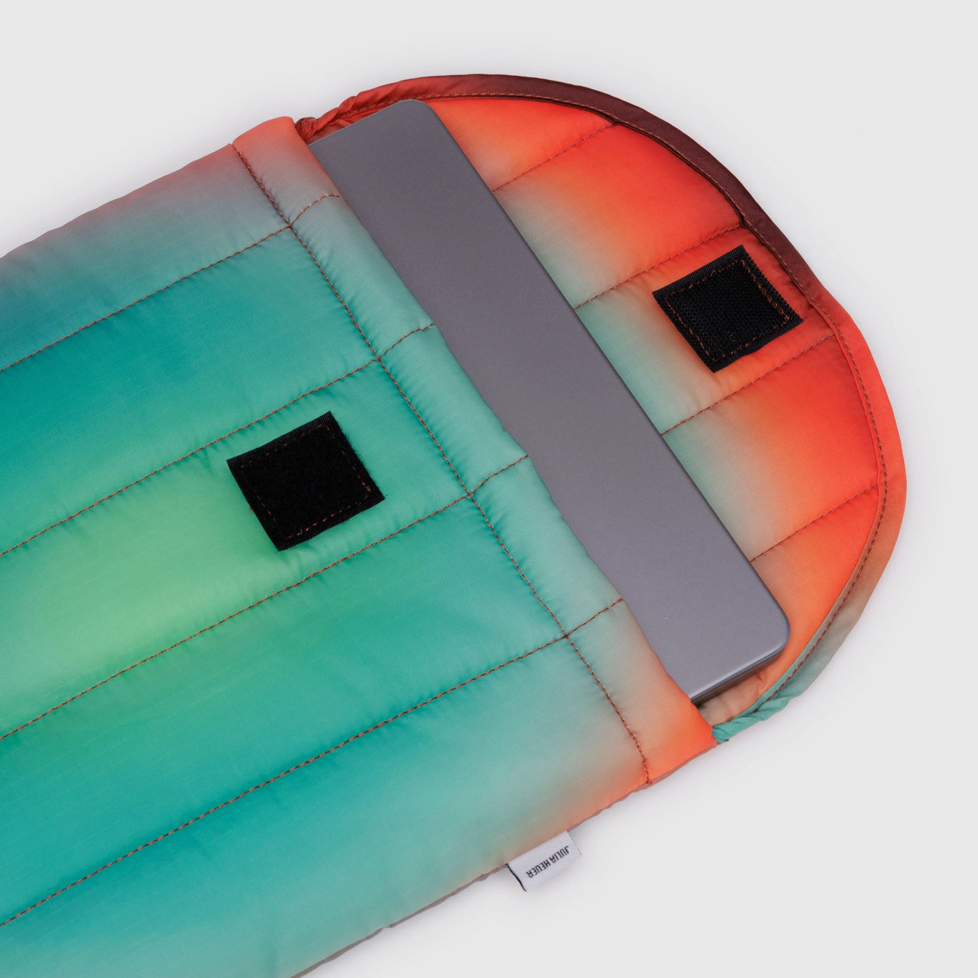 A laptop partially nestled in a colorful, padded sleeve featuring gradient shades of orange, red, teal, and green. This Baggu Puffy Laptop Sleeve 13"/14" - Rain boasts two sleek black Velcro strips on the flap—a true Gradient Masterpiece.