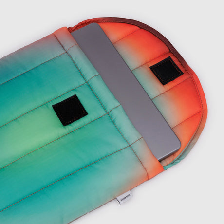 A laptop partially nestled in a colorful, padded sleeve featuring gradient shades of orange, red, teal, and green. This Baggu Puffy Laptop Sleeve 13"/14" - Rain boasts two sleek black Velcro strips on the flap—a true Gradient Masterpiece.