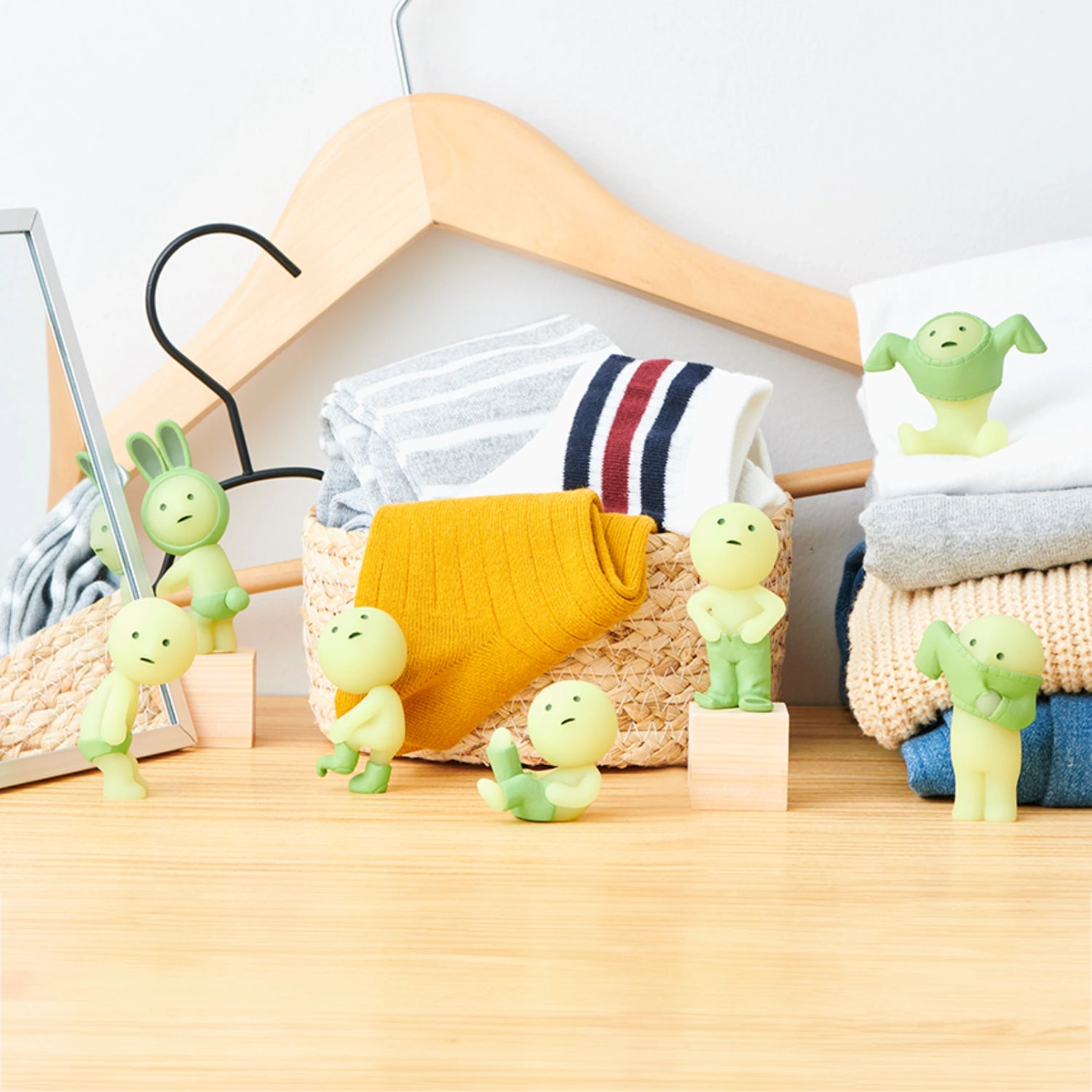 The Smiski Dressing Series - Blind Box features small green figurines positioned in various poses in front of a basket filled with clothes, hangers, and a folded mirror on a wooden surface. These captivating glow-in-the-dark figures by Smiski bring an enchanting touch to any display.