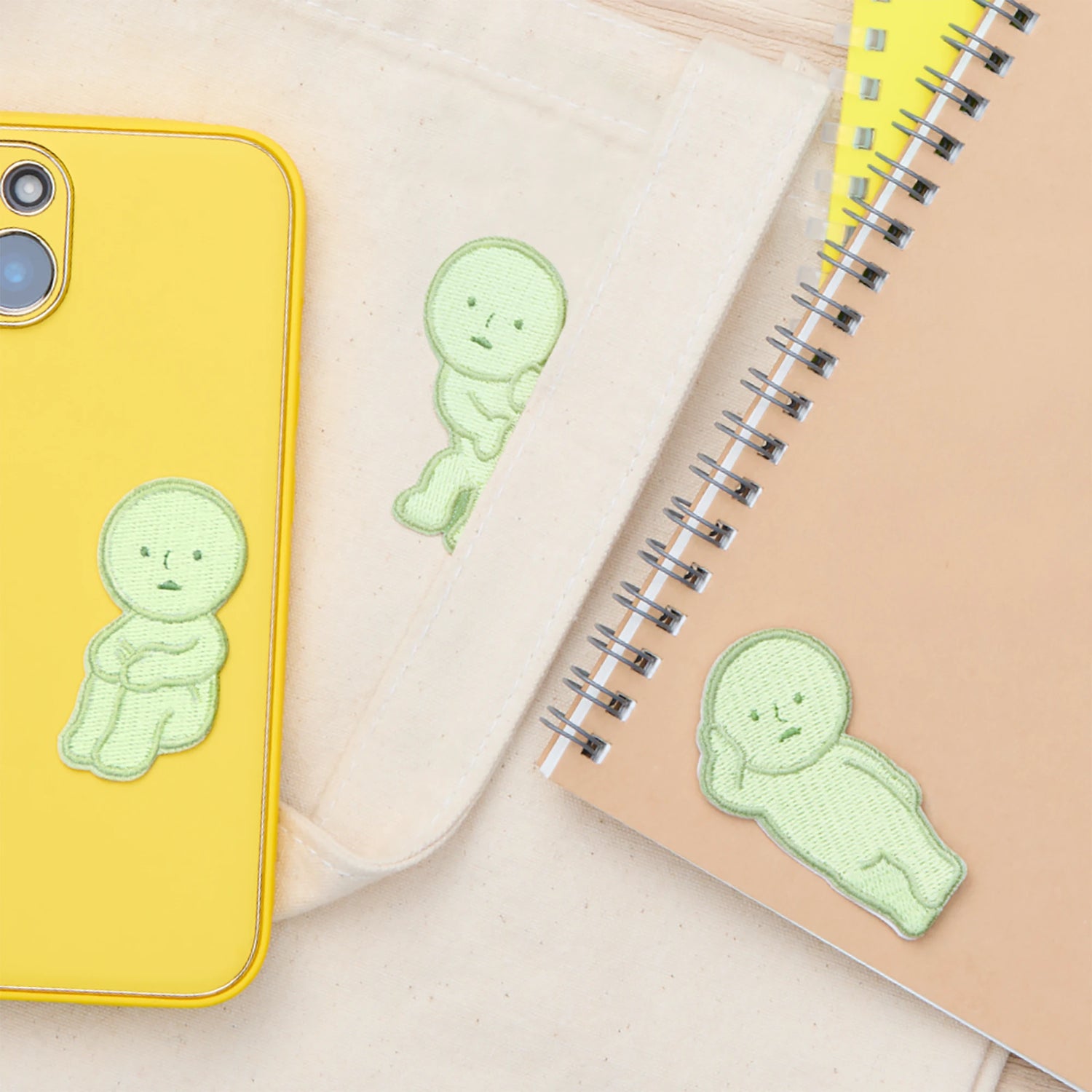 A yellow phone case, beige pouch, and spiral-bound notebook each feature a green sitting character Smiski Embroidery Sticker/Patch by Smiski, arranged on a neutral surface.