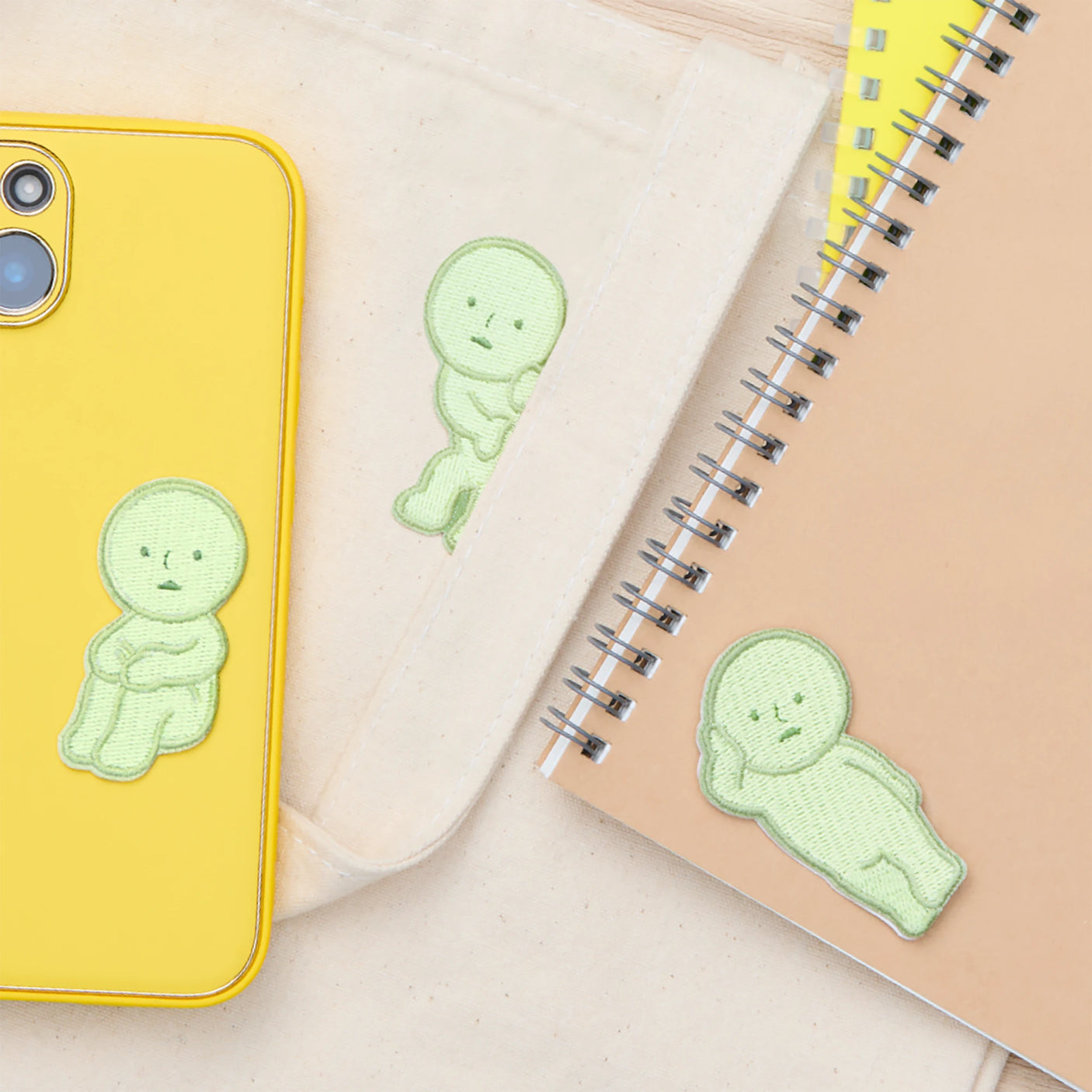 A yellow phone case, beige pouch, and spiral-bound notebook each feature a green sitting character Smiski Embroidery Sticker/Patch by Smiski, arranged on a neutral surface.