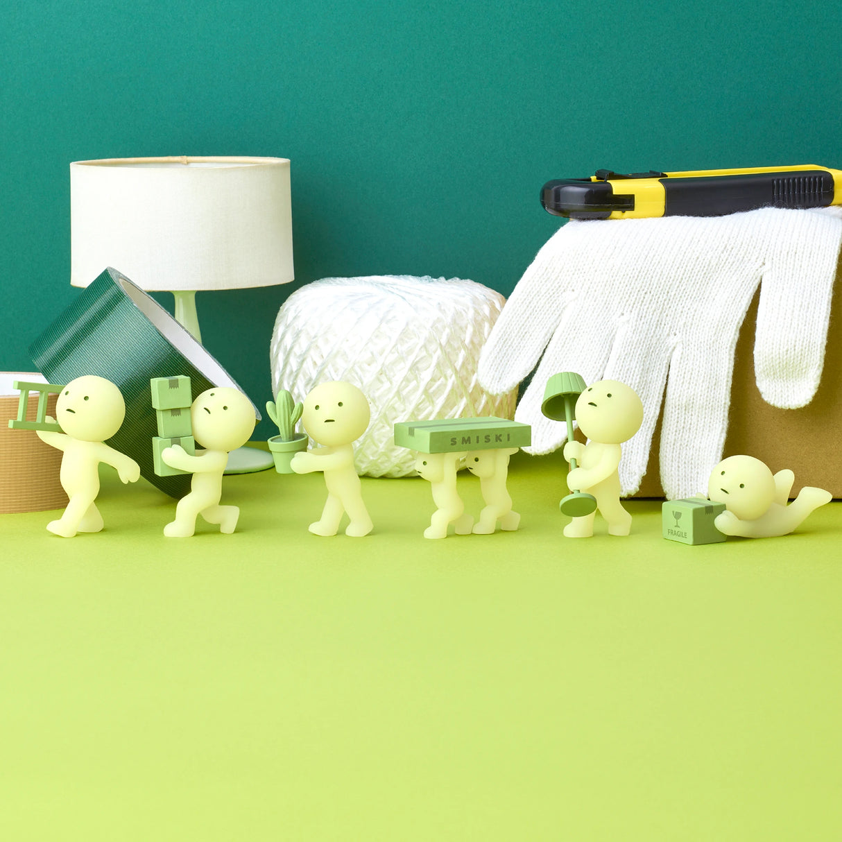 The Smiski Moving Series - Blind Box by Smiski features small green figurines arranged in a row, each holding items such as boxes and tape. Behind them, a surface adorned with a lamp, cutting tool, twine, and glove adds to the scene. These glow-in-the-dark collectible toys bring a whimsical touch to any setting.