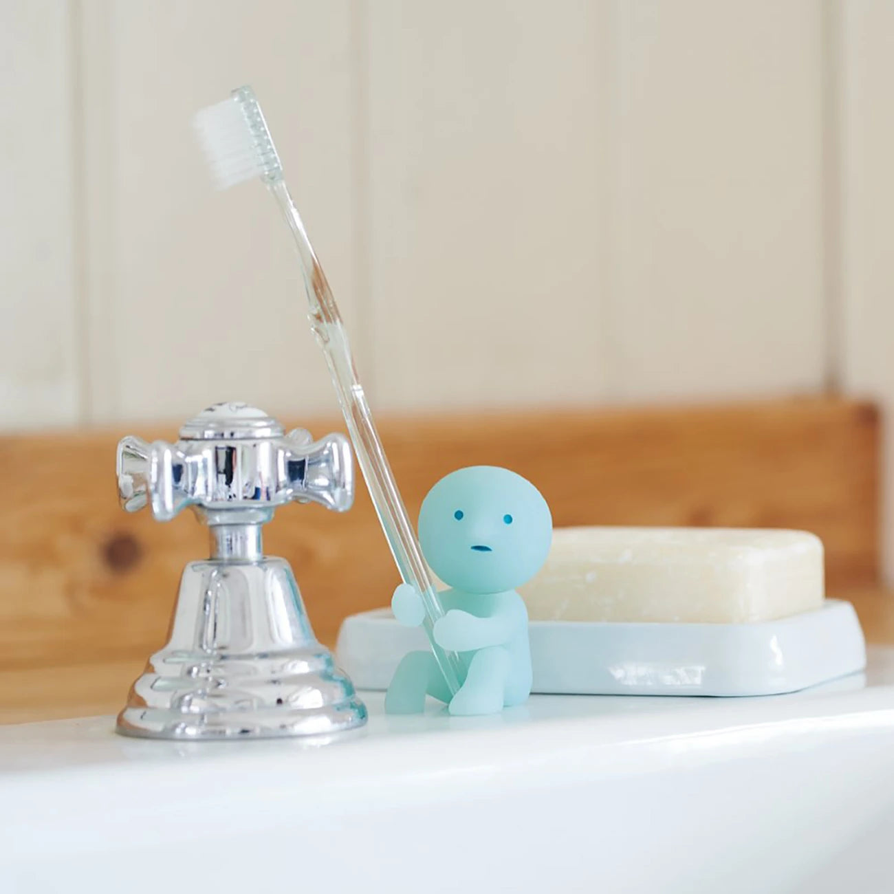 A Smiski Toothbrush Holder - Protecting Figure, your glow-in-the-dark guardian, perches next to a metallic tap handle and a bar of soap on a white bathroom surface.