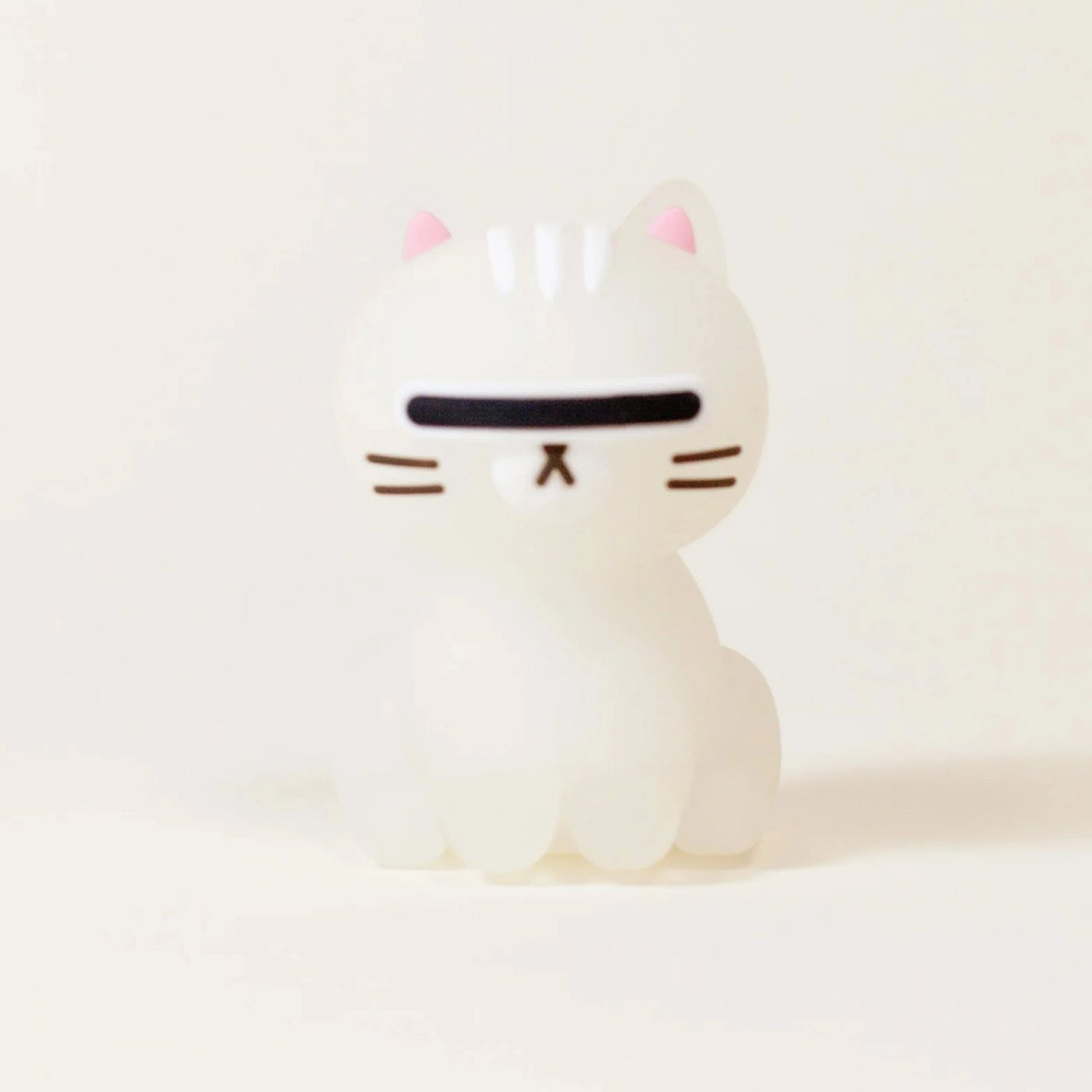A small white cat figurine from the Smoko Astrocats Blind Box by Smoko, featuring a minimalist design, black visor-like eyes, pink ears, and brown whiskers on a plain light background.