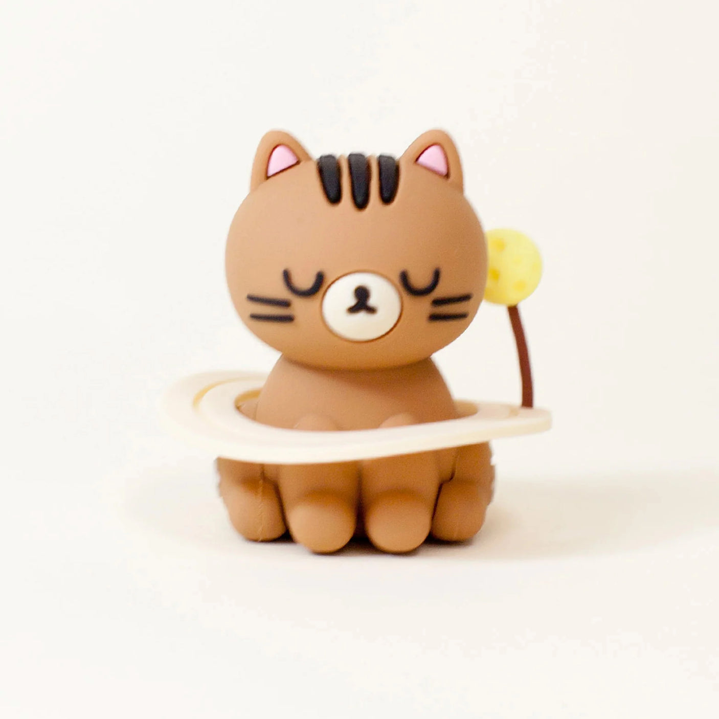 A small brown Smoko Astrocats Blind Box cartoon cat figurine with closed eyes sits in a cream ring with a yellow ball, set against a white background—an adorable collectible from Smoko.