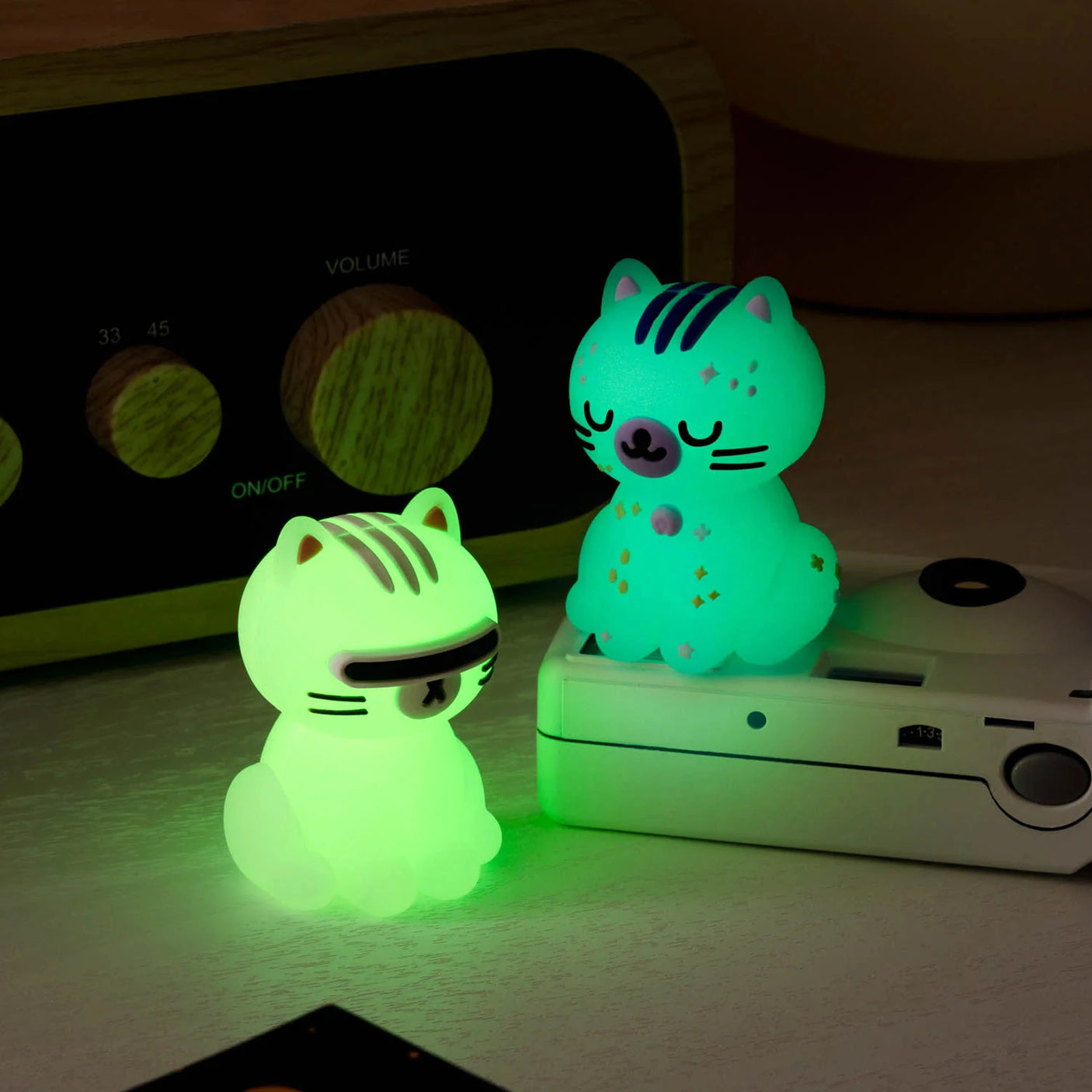 Two glowing Smoko Astrocats Blind Box figures—a striped, closed-eye cat and a visor-wearing cat—sit on a desk beside a retro radio and camera, enhancing the appeal of these collectible vinyl figures by Smoko.