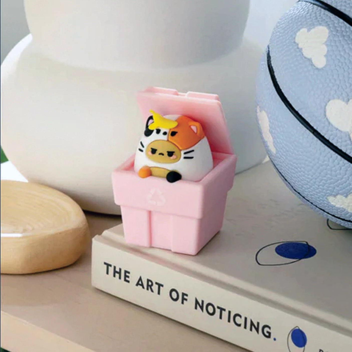 A small toy resembling an egg with tiger features is in a pink box from the Smoko Tayto Cat Blind Box collection. Nearby, "The Art of Noticing" book and a blue ball with clouds add charm to the scene.