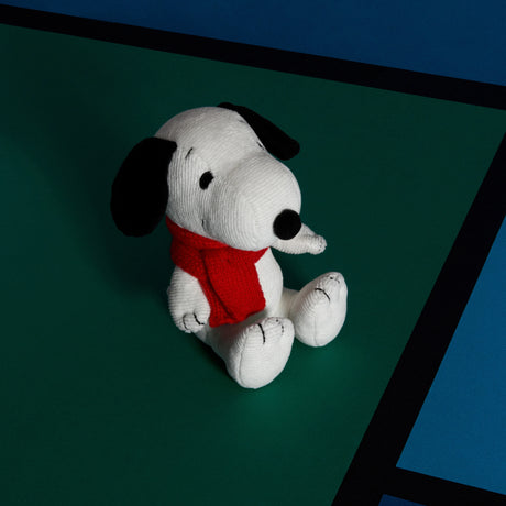 The Snoopy Scarf Plush - 17cm by Snoopy features black ears and a red scarf, made from recycled polyester, and sits on a green and blue surface divided by black lines.