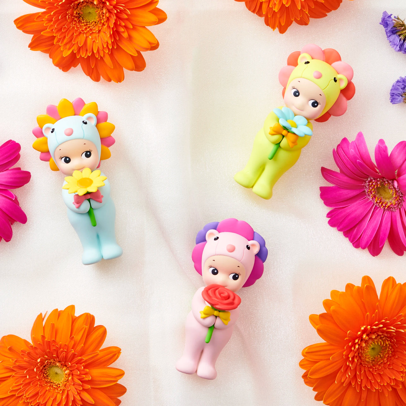 Three Sonny Angel Flower Gift Series figures from Sonny Angel in pastel animal costumes hold flowers among orange and pink gerbera daisies on a soft fabric—an ideal collectible or floral-themed gift.