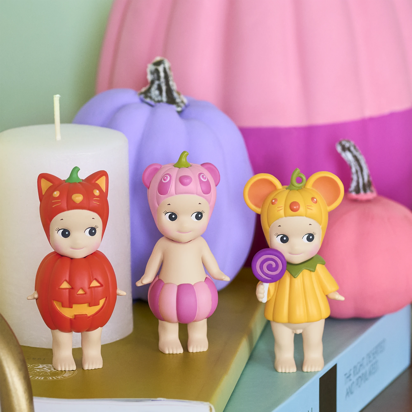 Six Sonny Angel Pumpkin Patch dolls, dressed in colorful pumpkin-themed costumes, stand on stacked books with pastel pumpkins and a white candle nearby. This limited edition sealed set by Sonny Angel adds festive charm to any display.