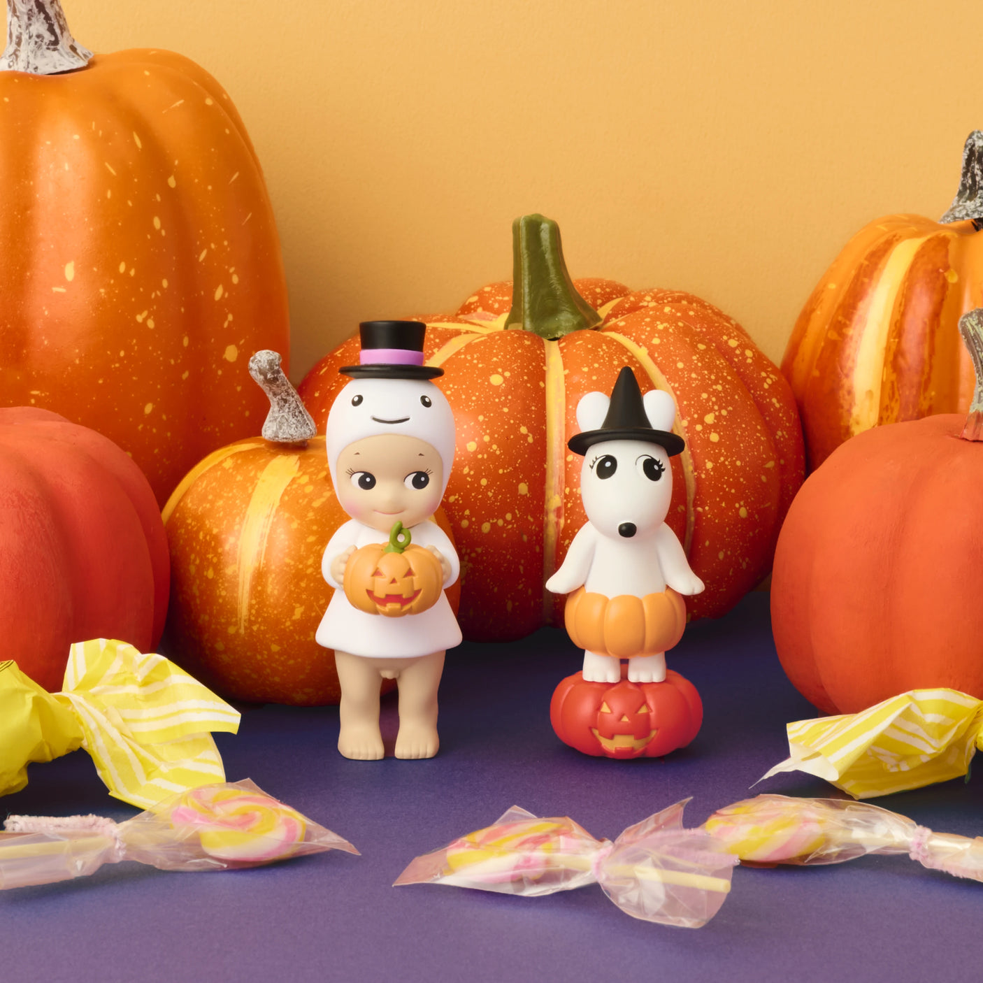 Two Sonny Angel Pumpkin Patch Blind Box - Limited Edition dolls stand among pumpkins and candies, set against an orange and purple background. One wears a ghost costume, while the other is dressed as a witch. Brand: Sonny Angel.