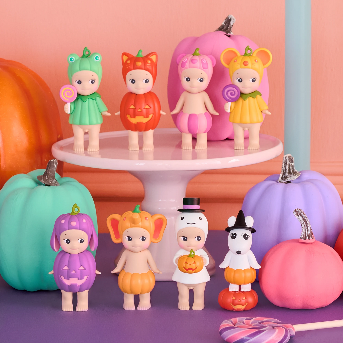 Eight limited edition Sonny Angel Pumpkin Patch Blind Box figures by Sonny Angel stand on a white cake stand, surrounded by pastel pumpkins and lollipops on a purple surface, celebrating Halloween.