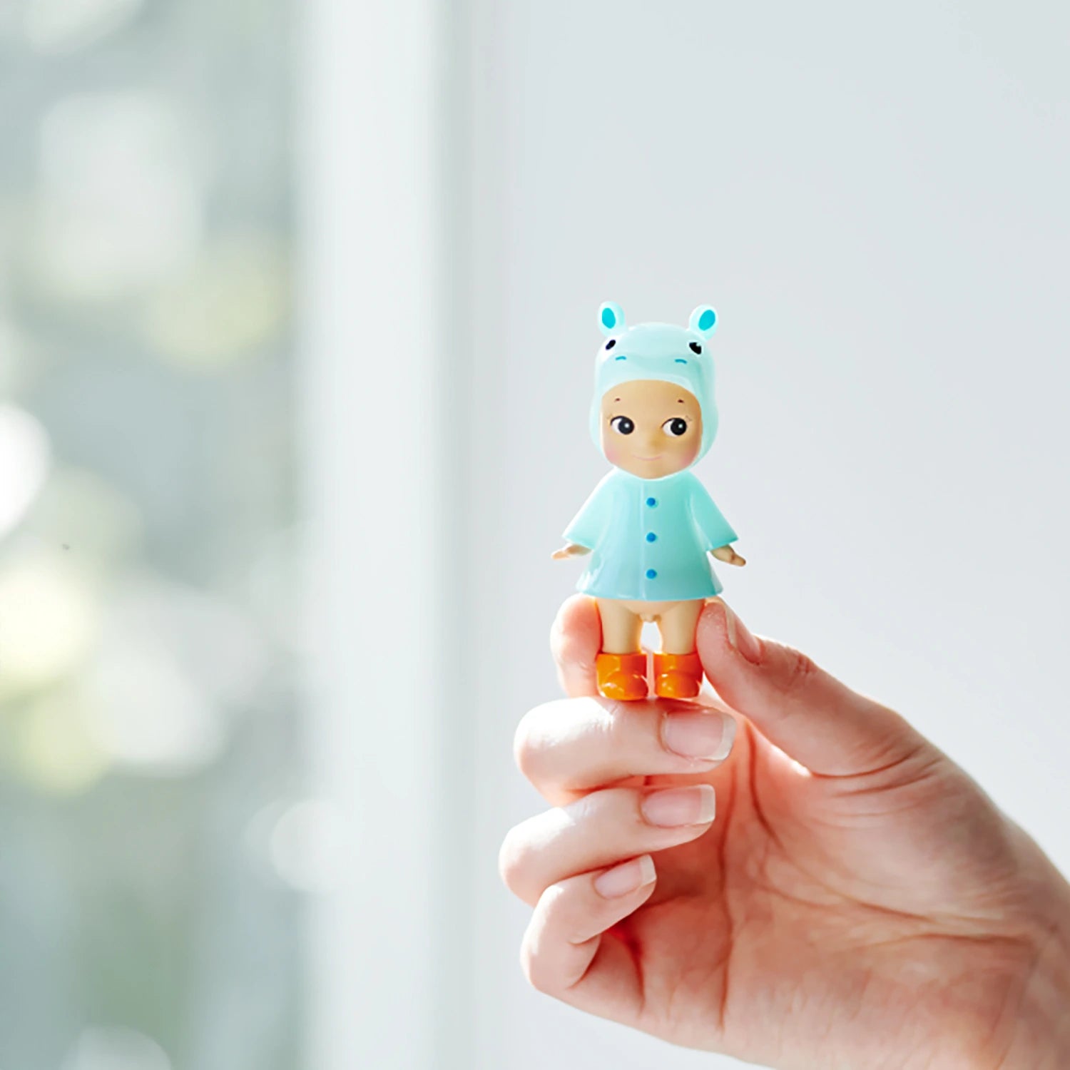 A hand holds a Sonny Angel Rainy Day Blind Box - Limited Edition doll by Sonny Angel, dressed in a light blue animal-themed hooded outfit and orange boots, set against a softly blurred background.