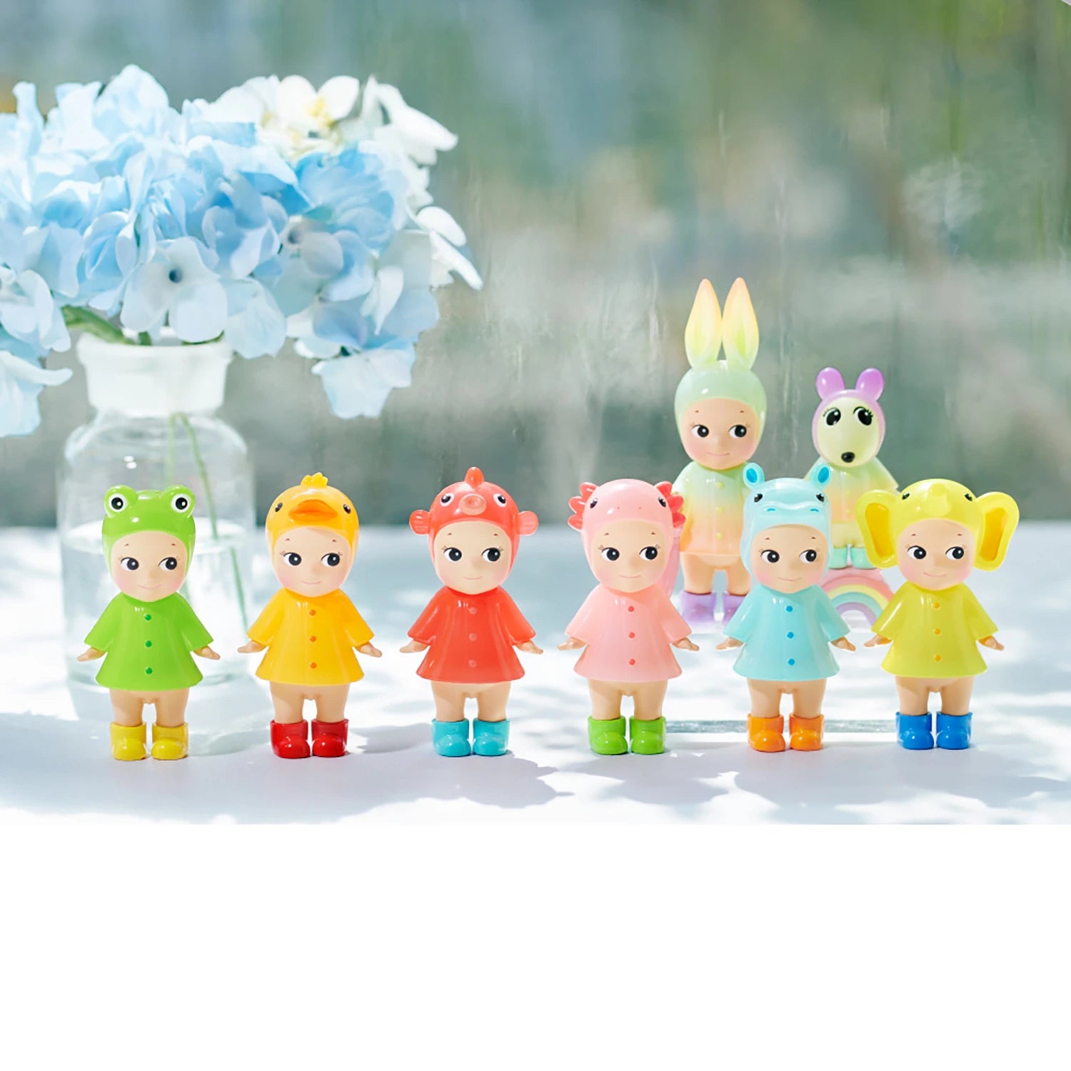 Seven Sonny Angel Rainy Day Blind Box - Limited Edition dolls from Sonny Angel, each in cute animal-themed outfits, stand in a row on a white surface with blue hydrangeas and another Blind Box doll blurred in the background.