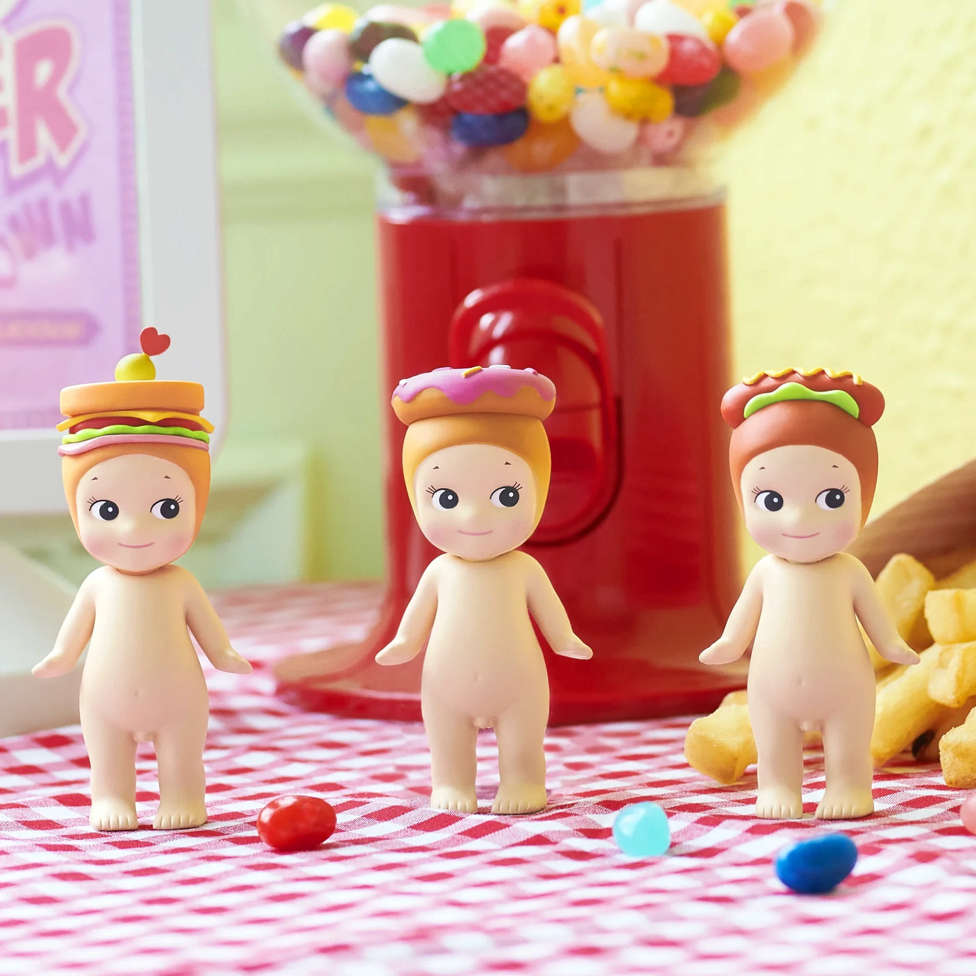 Three Sonny Angel Snack Series - Blind Box mini-figures, each wearing a food-themed hat—burger, donut, and hotdog—stand on a checkered tablecloth with jellybeans and a candy machine in the background. Brand: Sonny Angel.