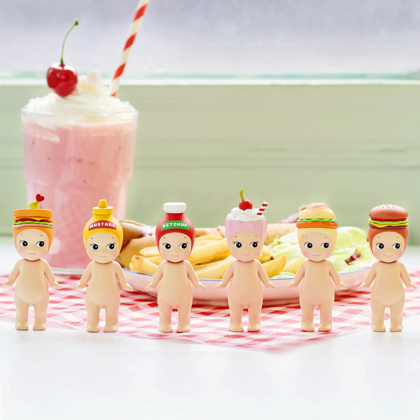 Six Sonny Angel Snack Series - Blind Box mini-figures, each with a cute snack-themed hat, stand in a row on a pink checkered cloth in front of a milkshake and plates of food. A perfect collectible from Sonny Angel for blind box fans!.