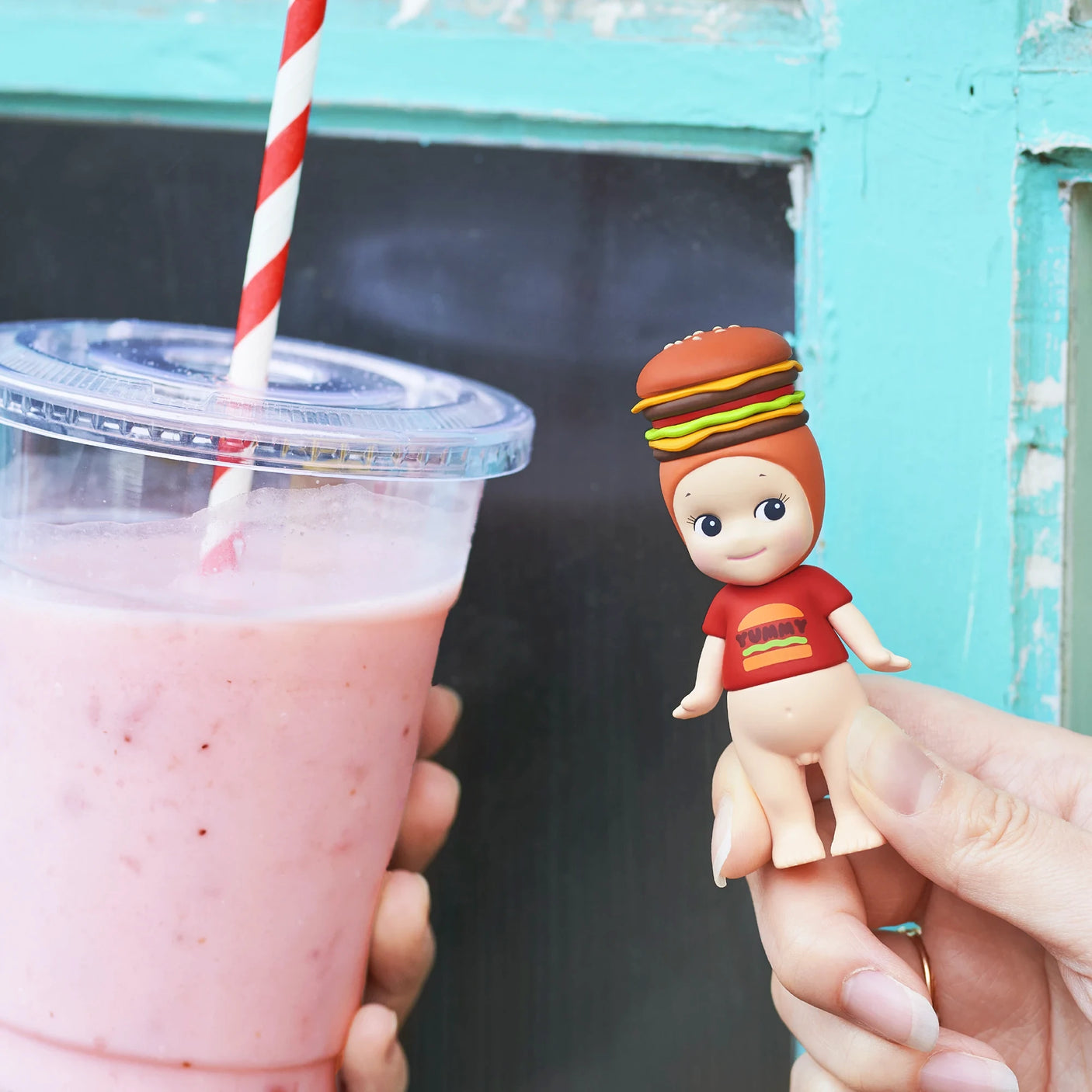 A hand holds a pink smoothie with a striped straw beside a Sonny Angel Snack Series - Blind Box mini-figure by Sonny Angel, wearing a hamburger hat and red shirt, set against a blue-green window frame.
